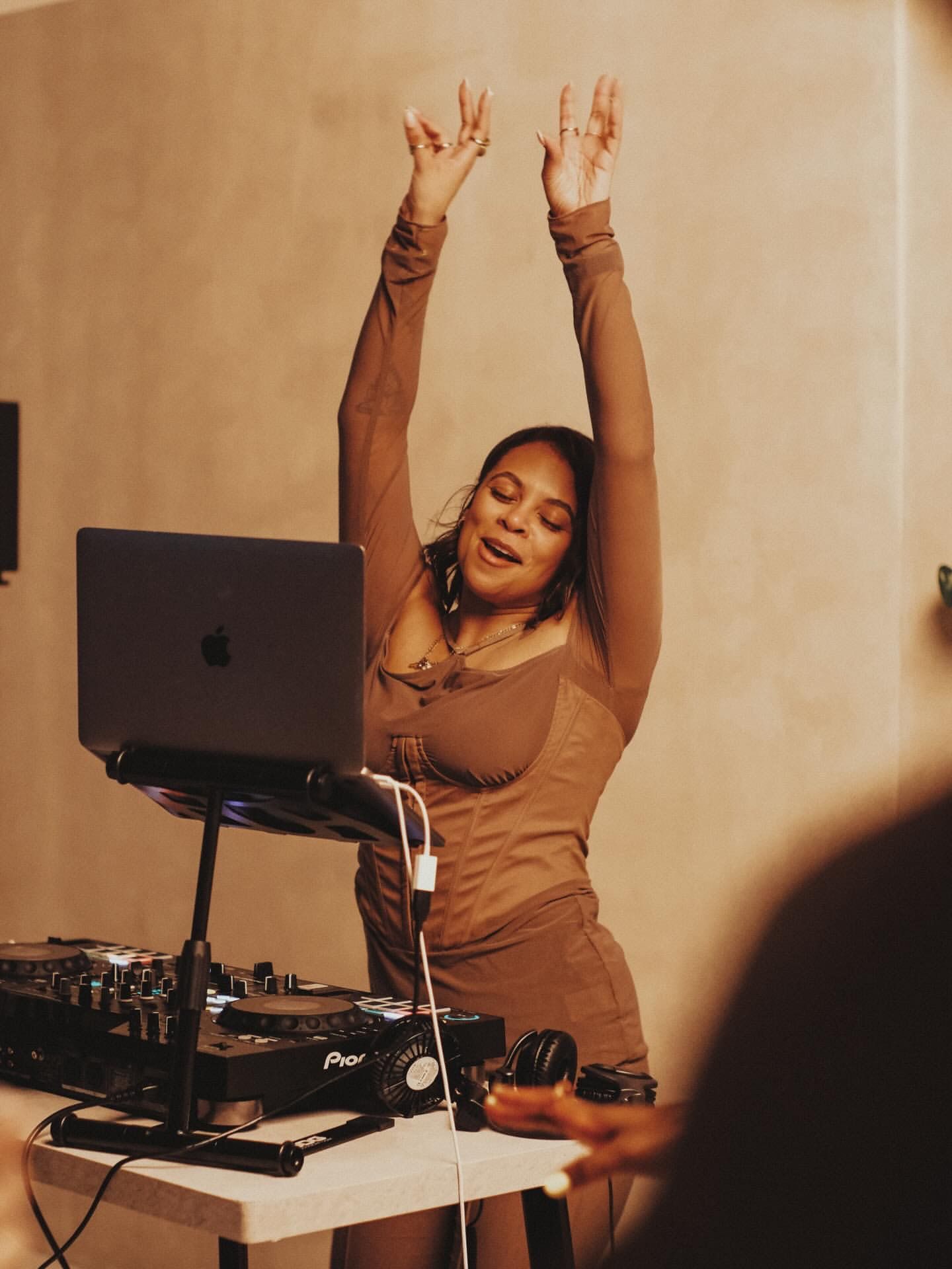 Female DJ with hands raised, playing music at a lively party in a warehouse venue.