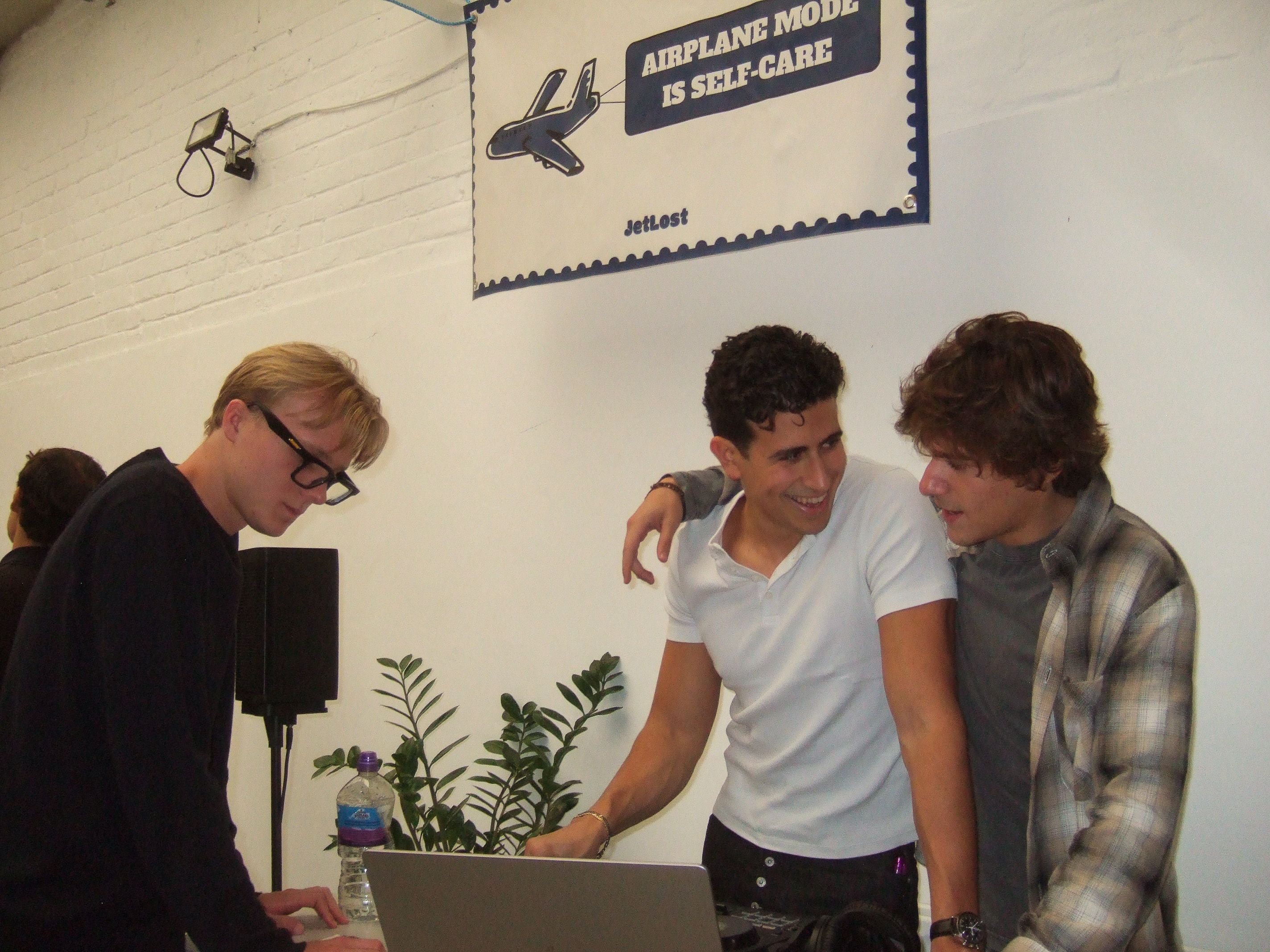 Three members of the JETLOST team share a laugh while setting up music equipment at Brixton Warehouse under a sign reading “Airplane Mode is Self-Care.”