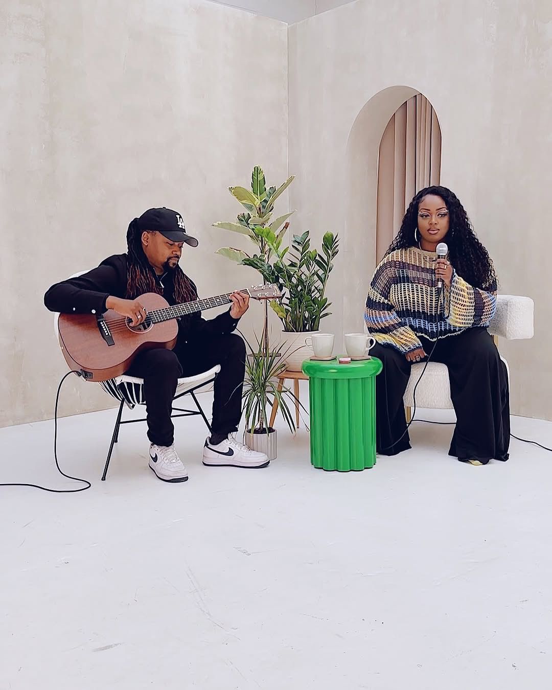 Unplugged performance by singer and guitarist in relaxed street style, set against limewashed art wall and bright green table in a modern studio.