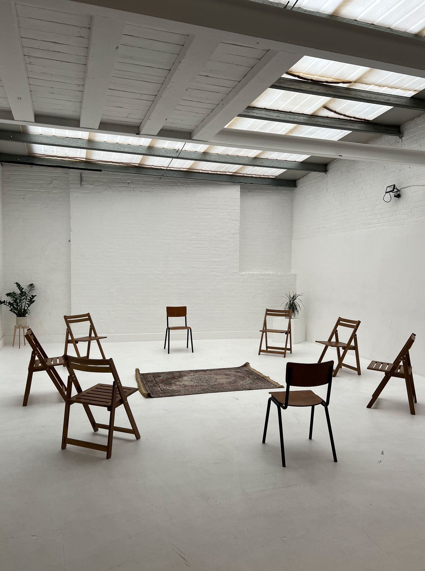 Alt Text / SEO Description "Minimalist studio at Brixton Warehouse set up for a life drawing class. Wooden chairs form a circle around a patterned rug in a bright white space with natural light from skylights."