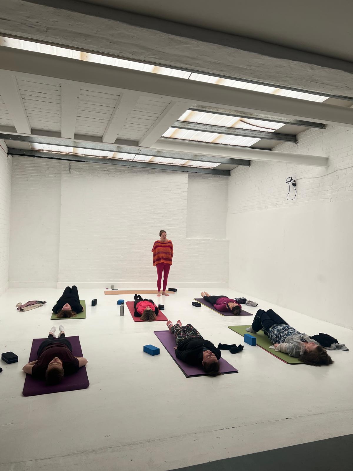 "Yoga class at Brixton Warehouse in a bright white studio. Participants lie on colorful mats in savasana relaxation pose while the instructor, dressed in a red and orange striped sweater, guides the session."