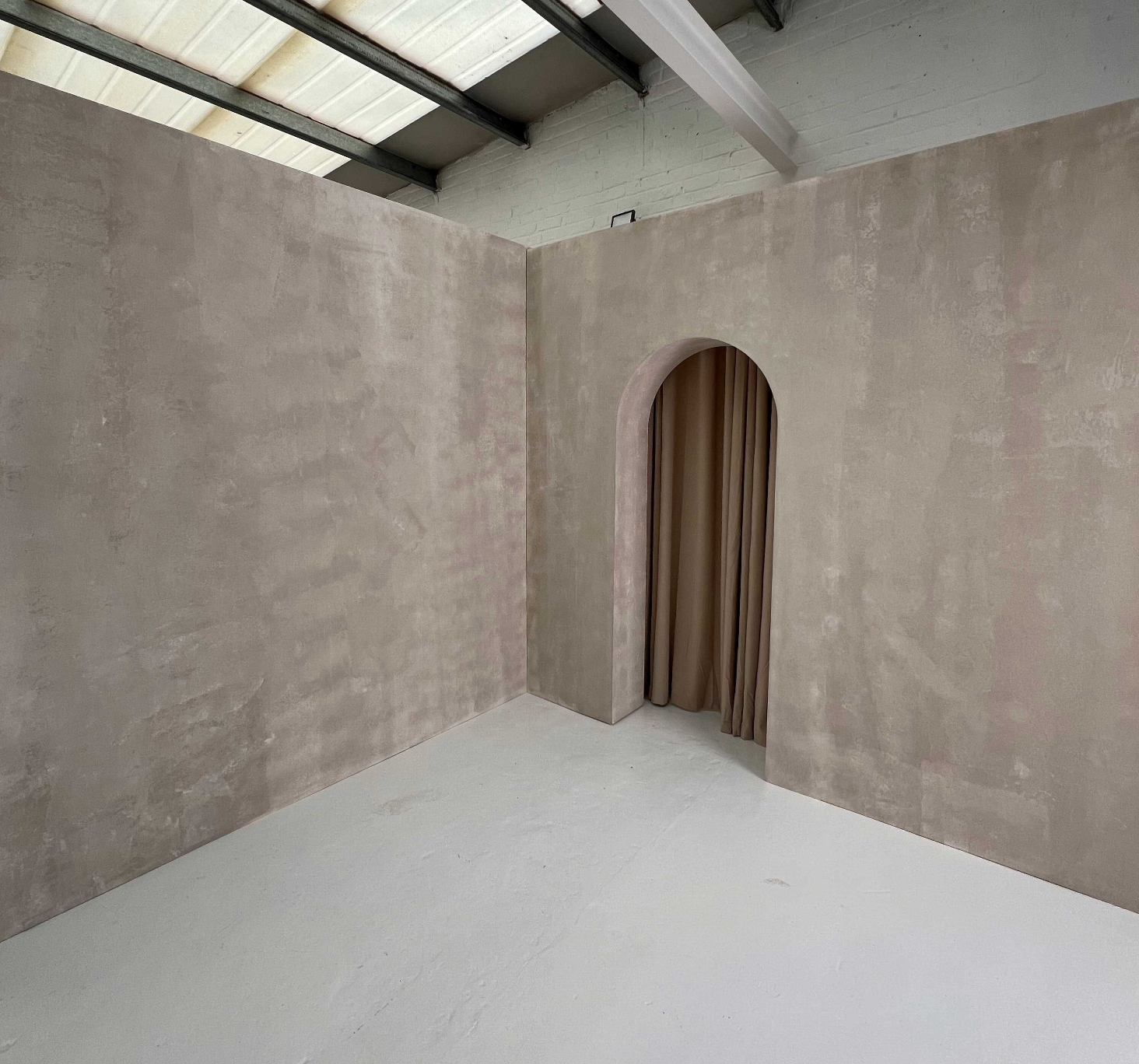 A minimalist set build featuring a textured, neutral-toned arched wall with a soft beige curtain. The space has an industrial aesthetic with exposed metal beams and a translucent ceiling, allowing natural light to filter in. The smooth, light-colored floor contrasts with the raw plastered walls, creating a modern, versatile backdrop. Perfect for photography, film shoots, and creative projects, this architectural design blends simplicity with elegance.