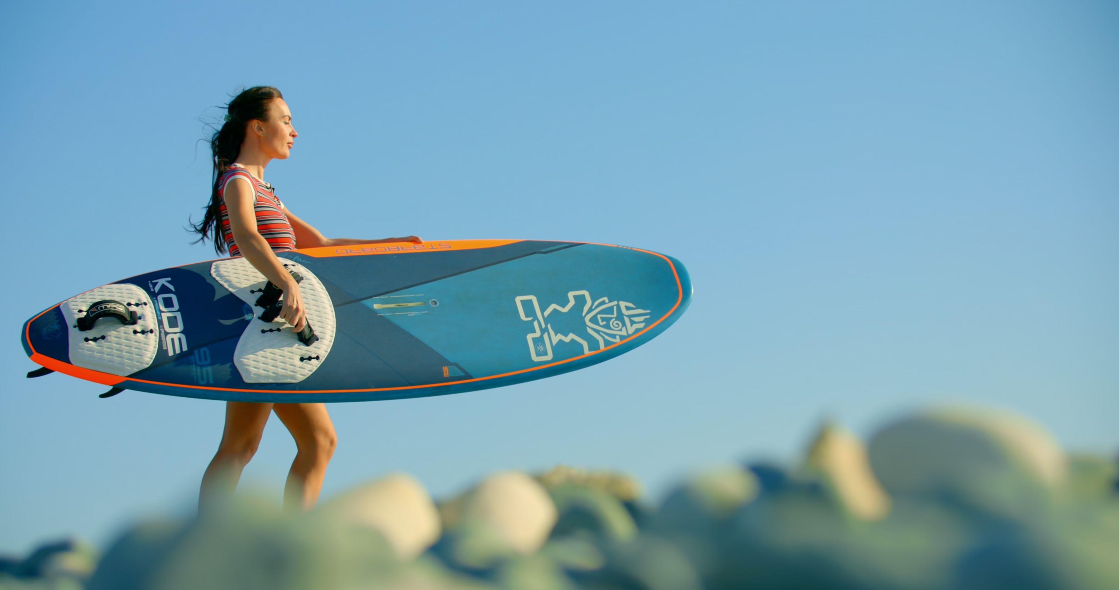 Woman carrying a windsurf board along the rocky coast
