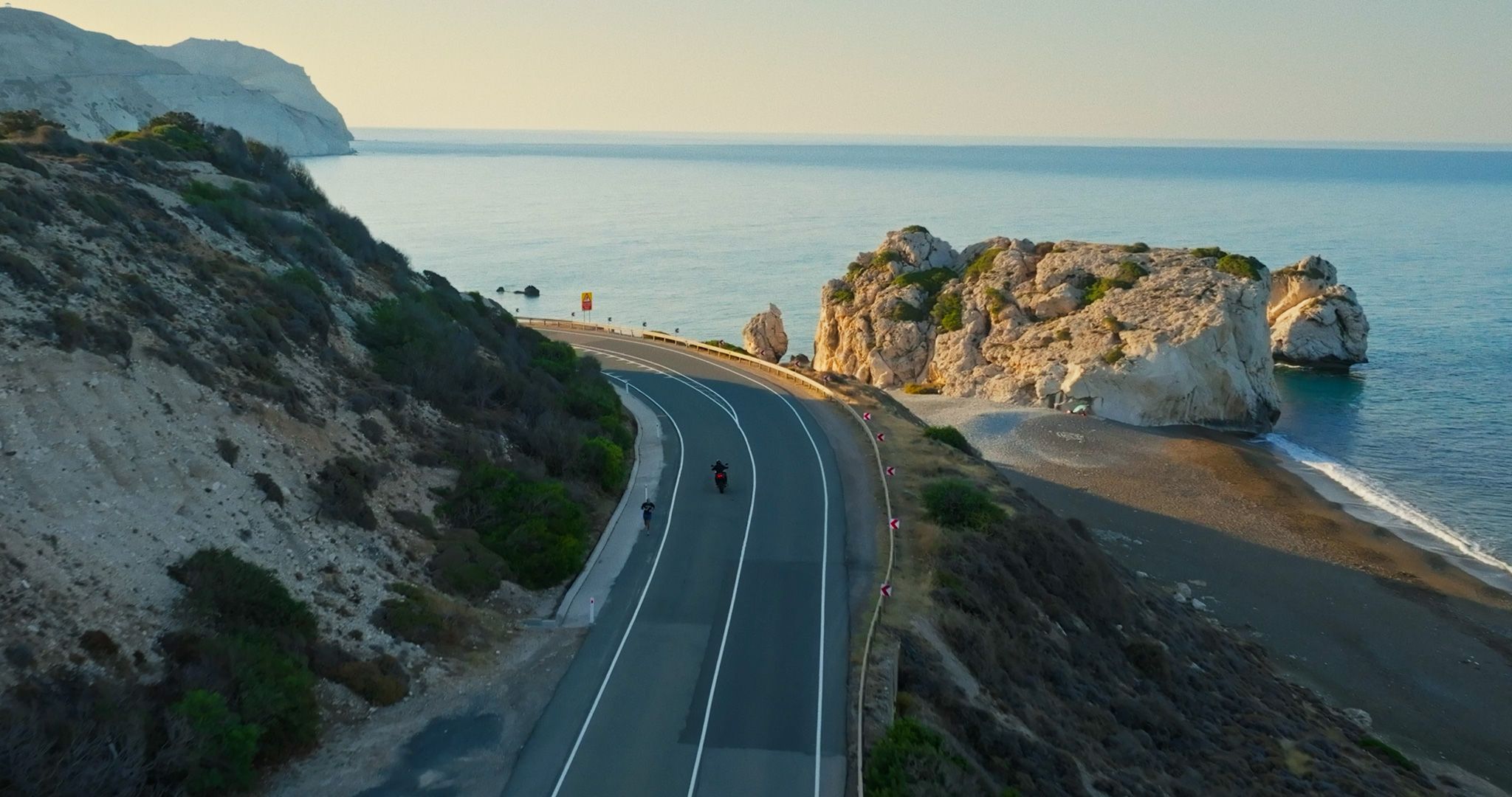 Aerial view of a coastal road winding between rocky cliffs and the sea near Paphos, with two riders on the road.