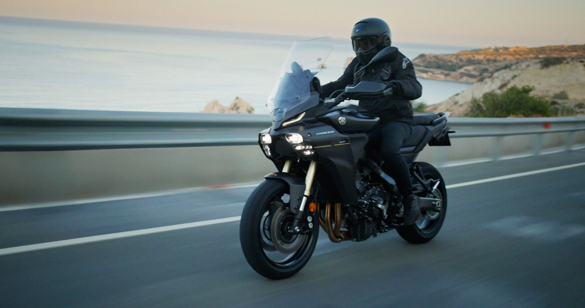 A motorcyclist riding along a coastal road with the sea and rocky landscape in the background.