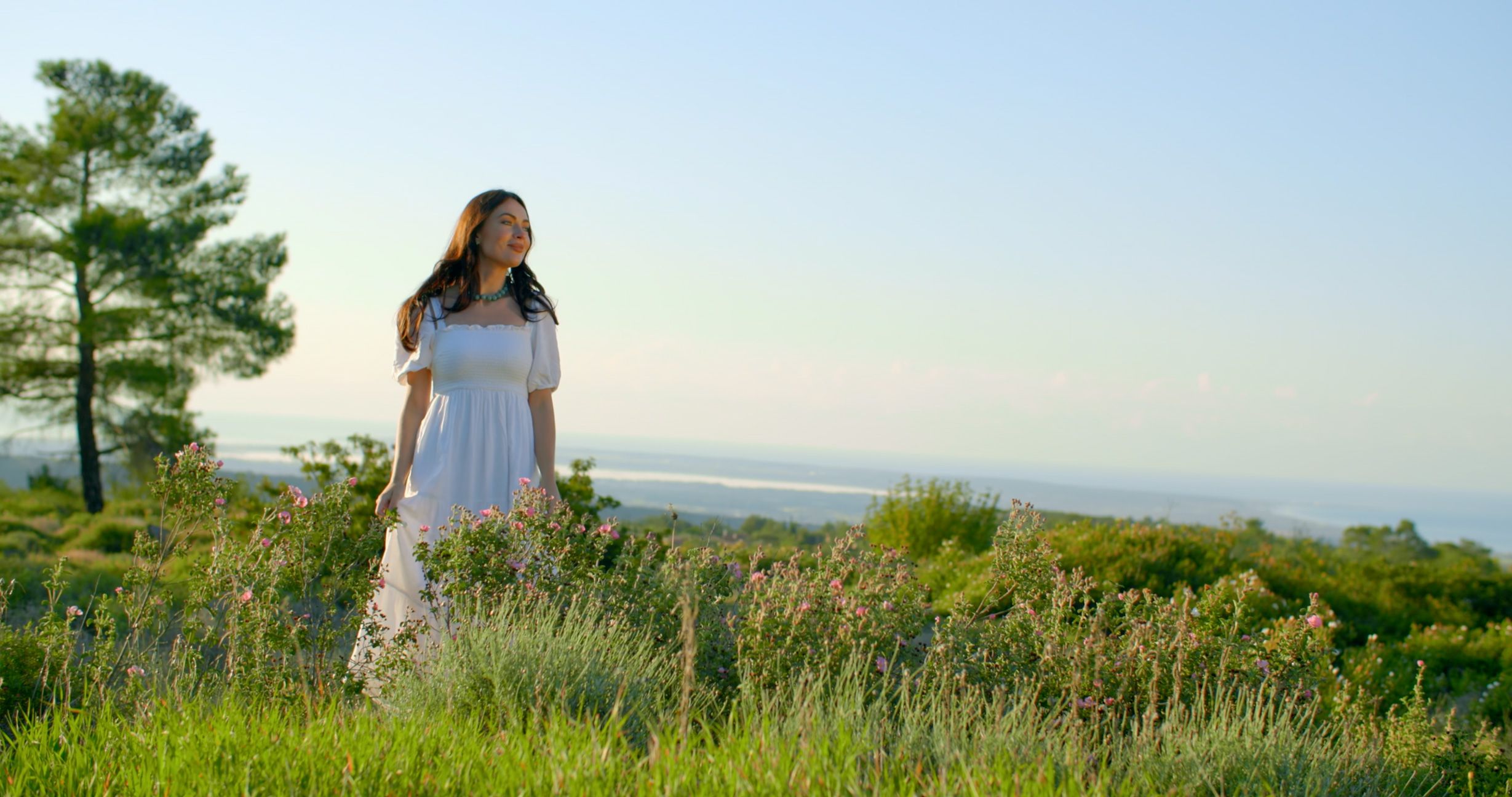 Woman in a white dress walking through wildflowers above the Cyprus coast
