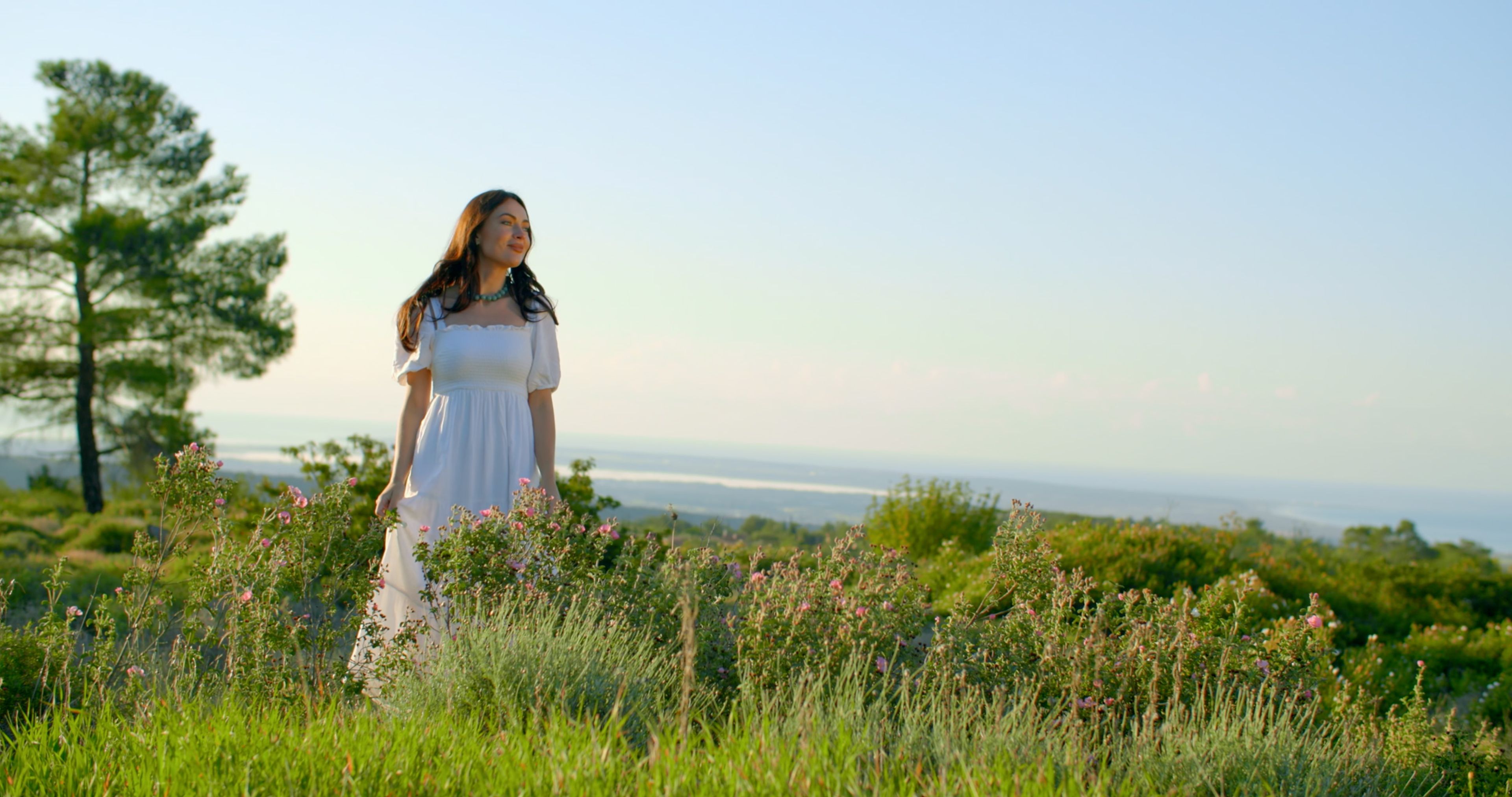 Woman in a white dress walking through wildflowers above the Cyprus coast