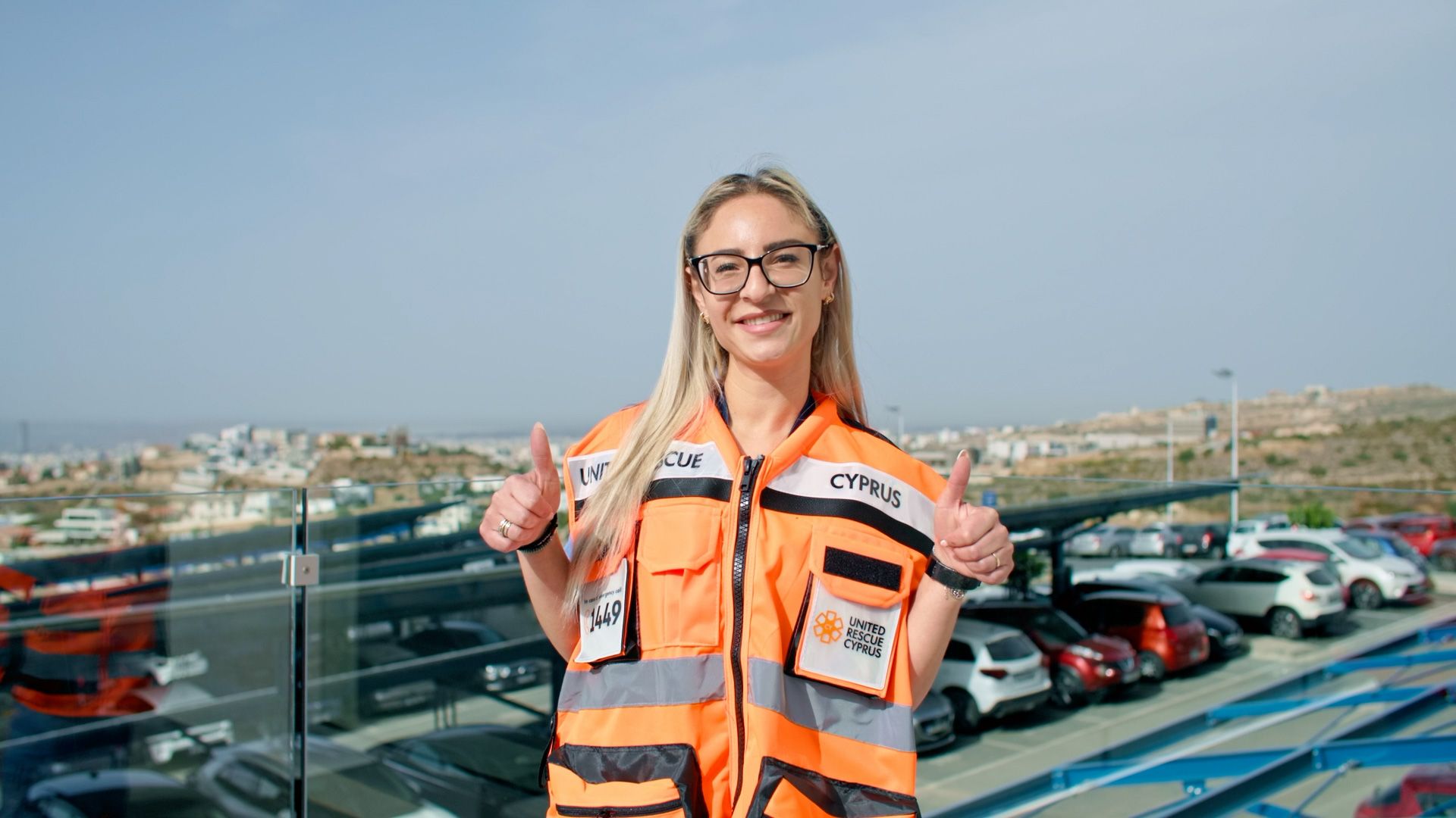 United Rescue Cyprus volunteer in an orange emergency vest giving a thumbs-up outdoors.
