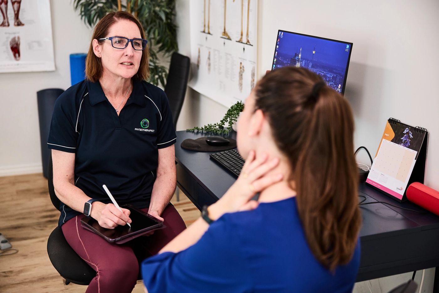 Physiotherapist taking notes on a tablet while talking with a patient in a consult room