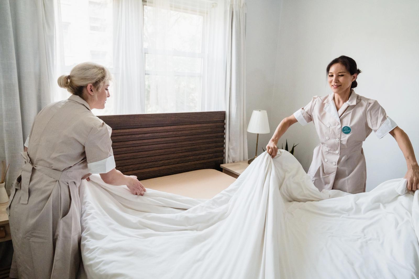 Two housekeepers changing white bed sheets in a bright guest room