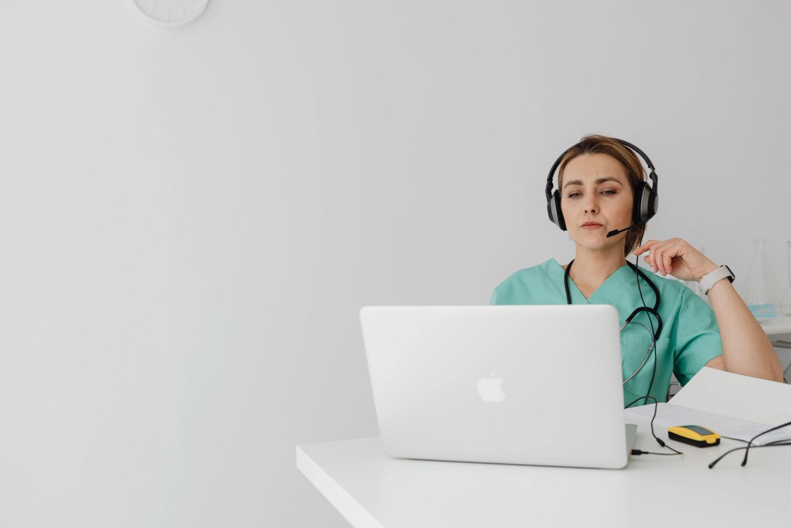 Healthcare professional in blue scrubs wearing a headset at a laptop in a medical office