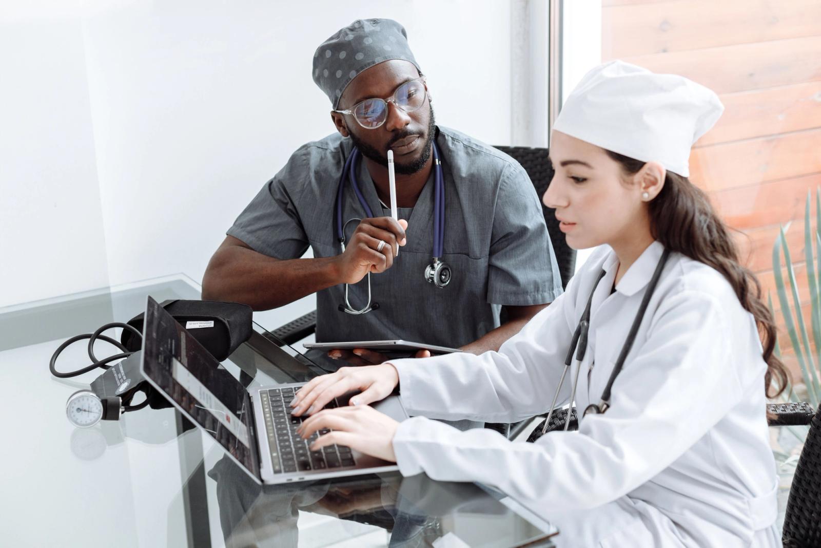 Two healthcare professionals in scrubs reviewing information on a laptop in a hospital office