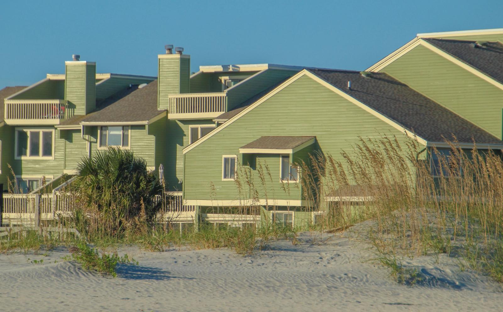Green beachfront vacation homes beside sand dunes under a blue sky
