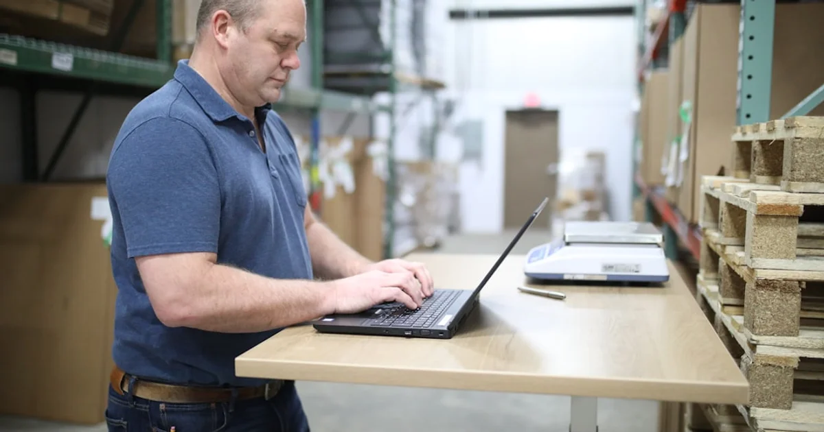 A man working on a laptop at a standing packing bench, a parcel scale beside him and warehouse racking behind