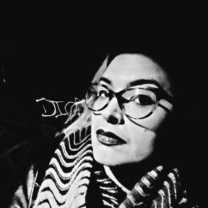A high-contrast black and white photo in which a genderqueer Palestinian person is leaning to the right and staring sidelong at the viewer against a dark background with graffiti. They are wearing black lipstick, large cat-eye glasses, a grey and black rippled scarf, and have long hair with a pale streak in front.