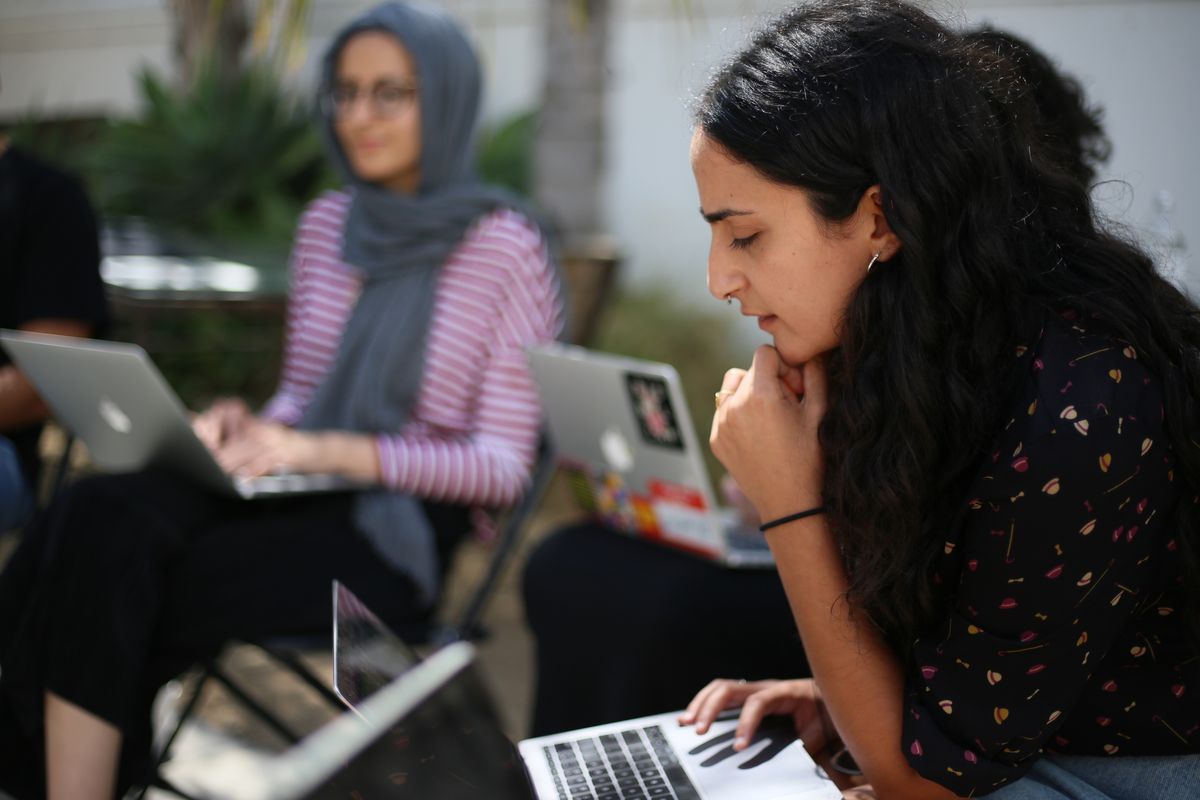 Outdoor photo by Shiyam Galyon (2019) of a RAWI member with long black hair and light brown skin wearing a black top with a small floral pattern reads from a computer with one hand on their chin. In the background, a light brown skinned person in a gray hijab and pink and white striped top and black pants looks on with a computer on their lap.