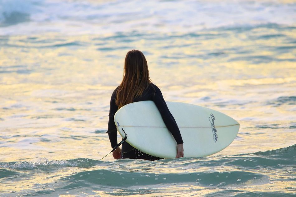 Person with long hair holding a white surfboard, looking out into the ocean.