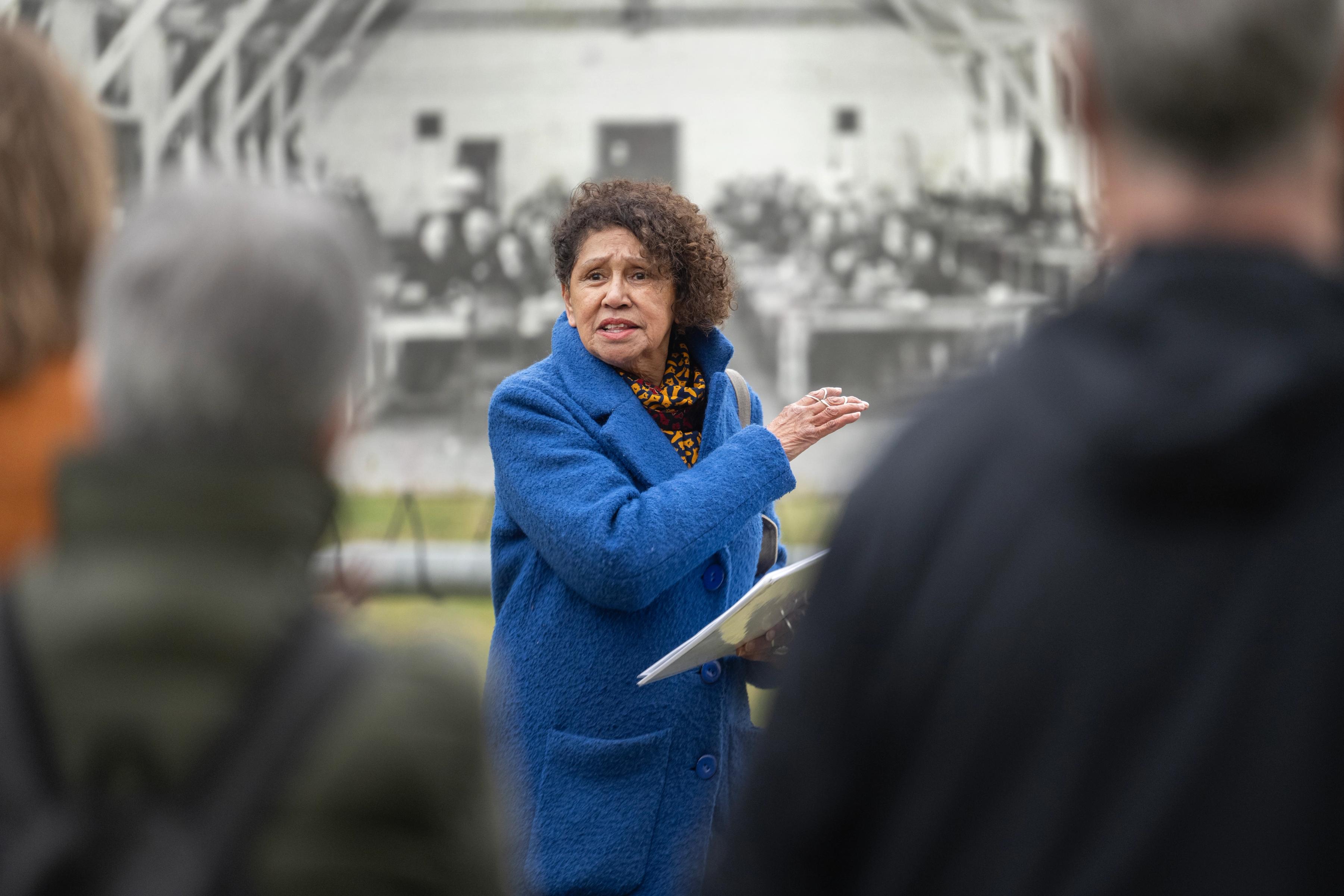 Molukse vrouw in felblauwe jas geeft rondleiding en staat voor een historische foto een wijst opzij, terwijl ze uitleg geeft aan de groep bezoekers.