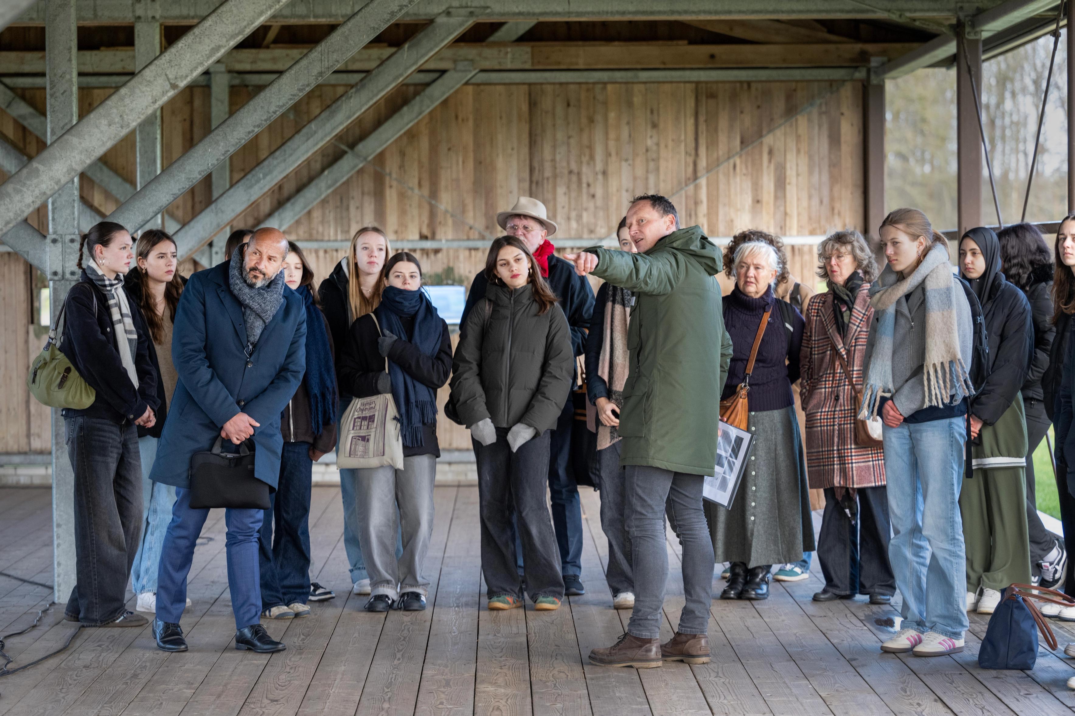Groep mensen kijkt naar rondleider die al wijzend het verhaal vertelt in een voormalige barak bij kamp westerbork