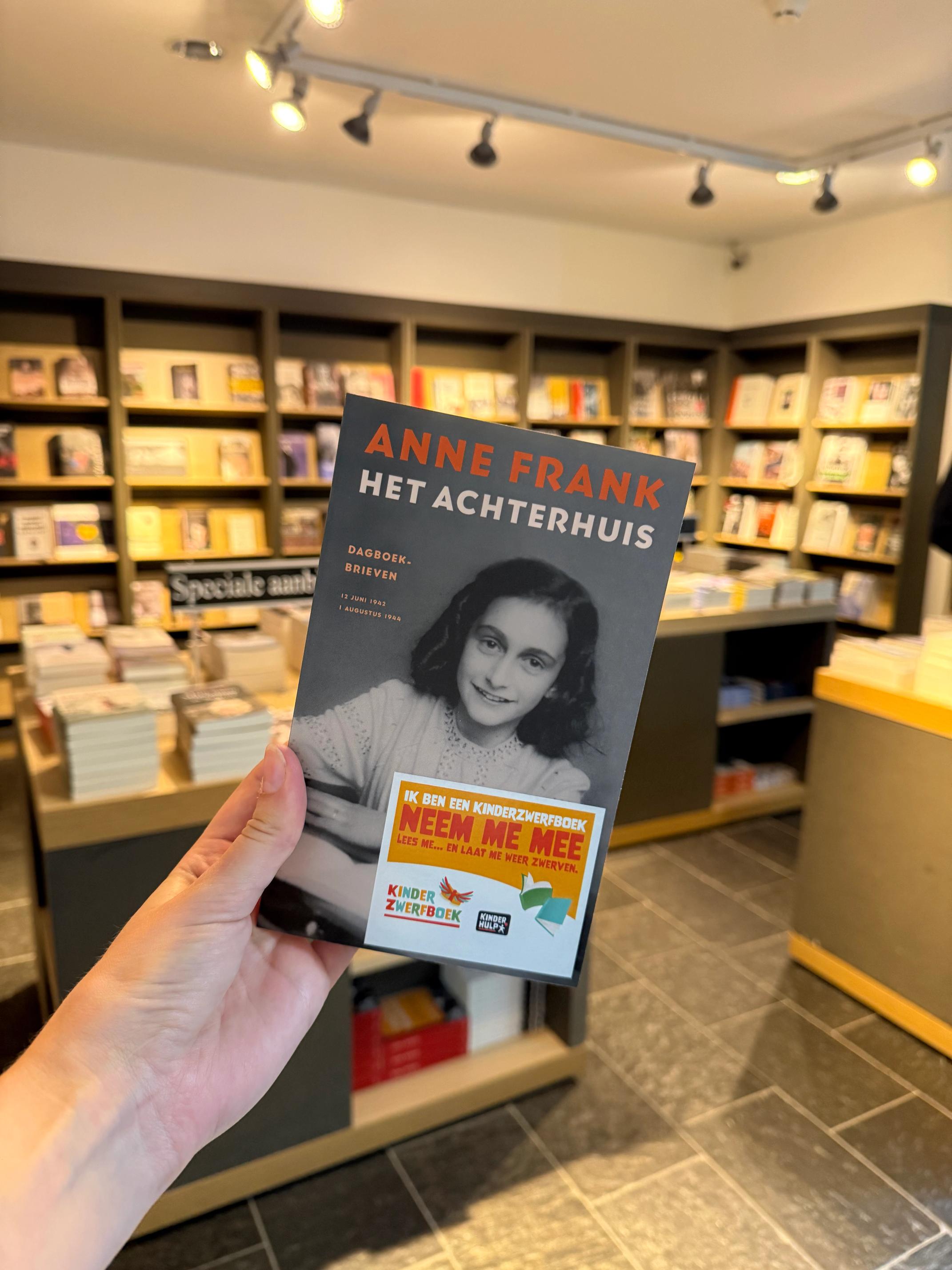 Hand houdt boek "Het Achterhuis" van Anne Frank vast, met op de achtergrond de boekenwinkel van Kamp Westerbork