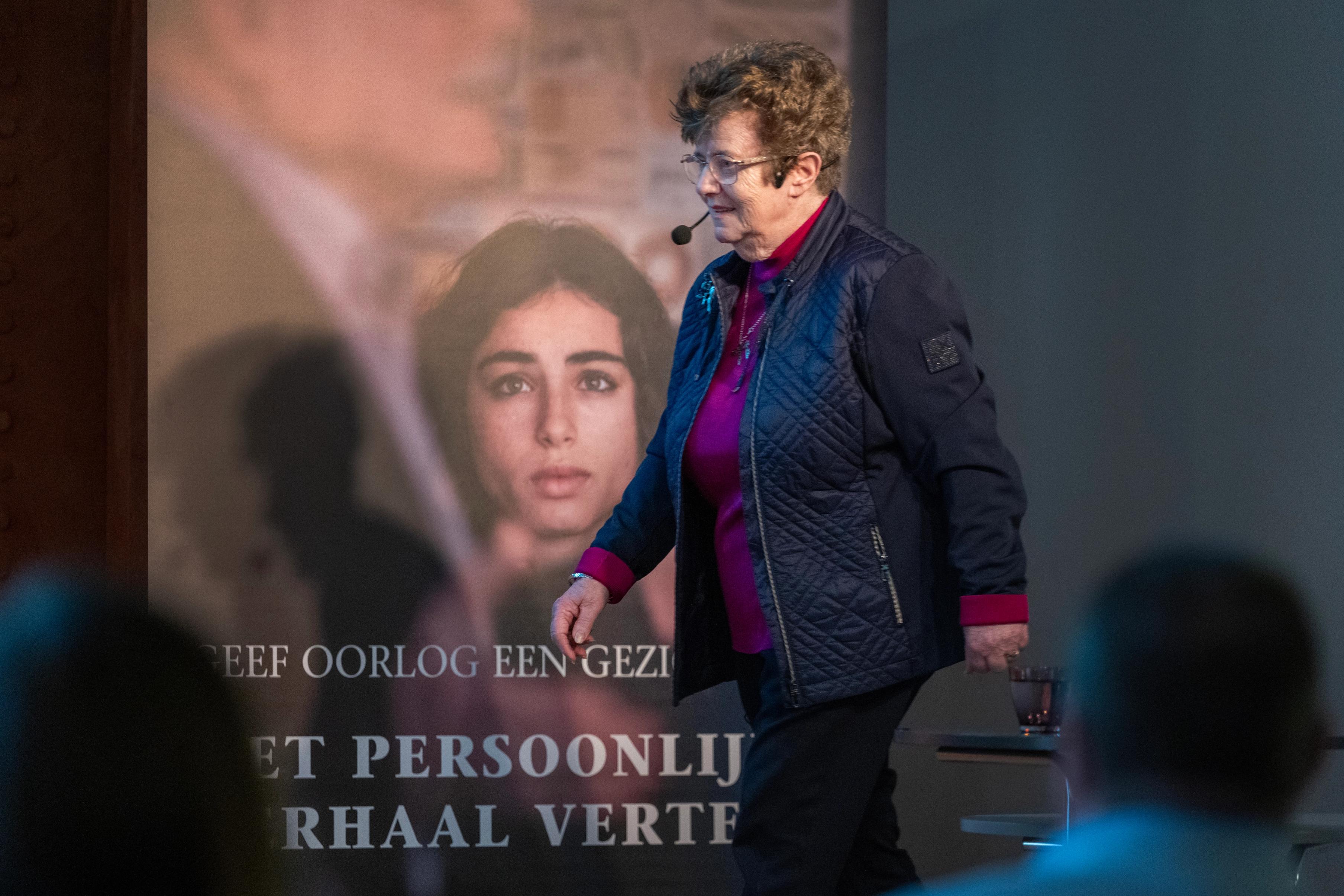 Oudere vrouw met een microfoon wandelt over een podium voor een publiek tijdens een ooggetuigenlezing