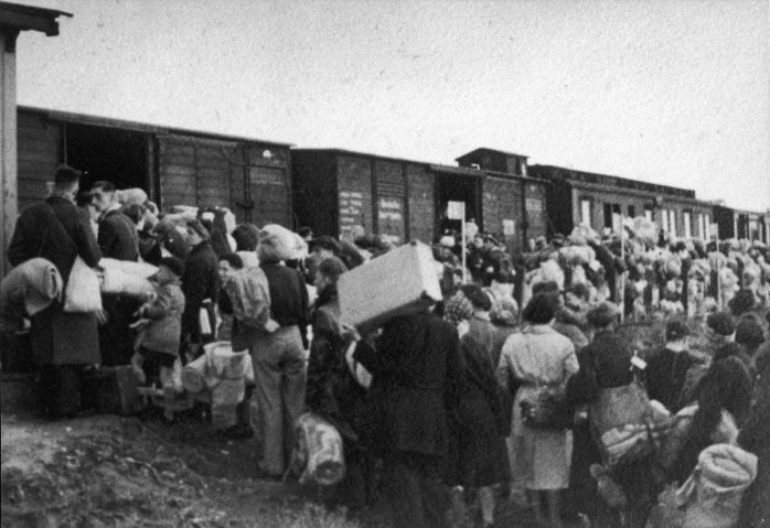 Oude zwart-wit collectiefoto van een groep mensen met koffers die staan te wachten voor de wagon op voormalig kampterrein Westerbork