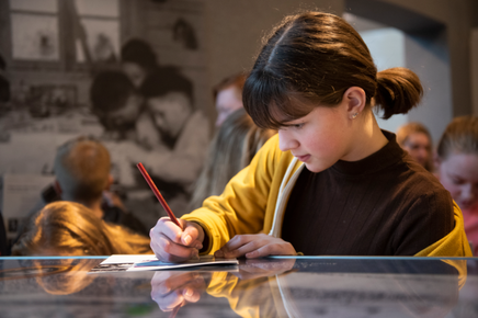 A girl in a yellow cardigan is writing with a pencil on a piece of paper in the museum at the Camp Westerbork Memorial Centre