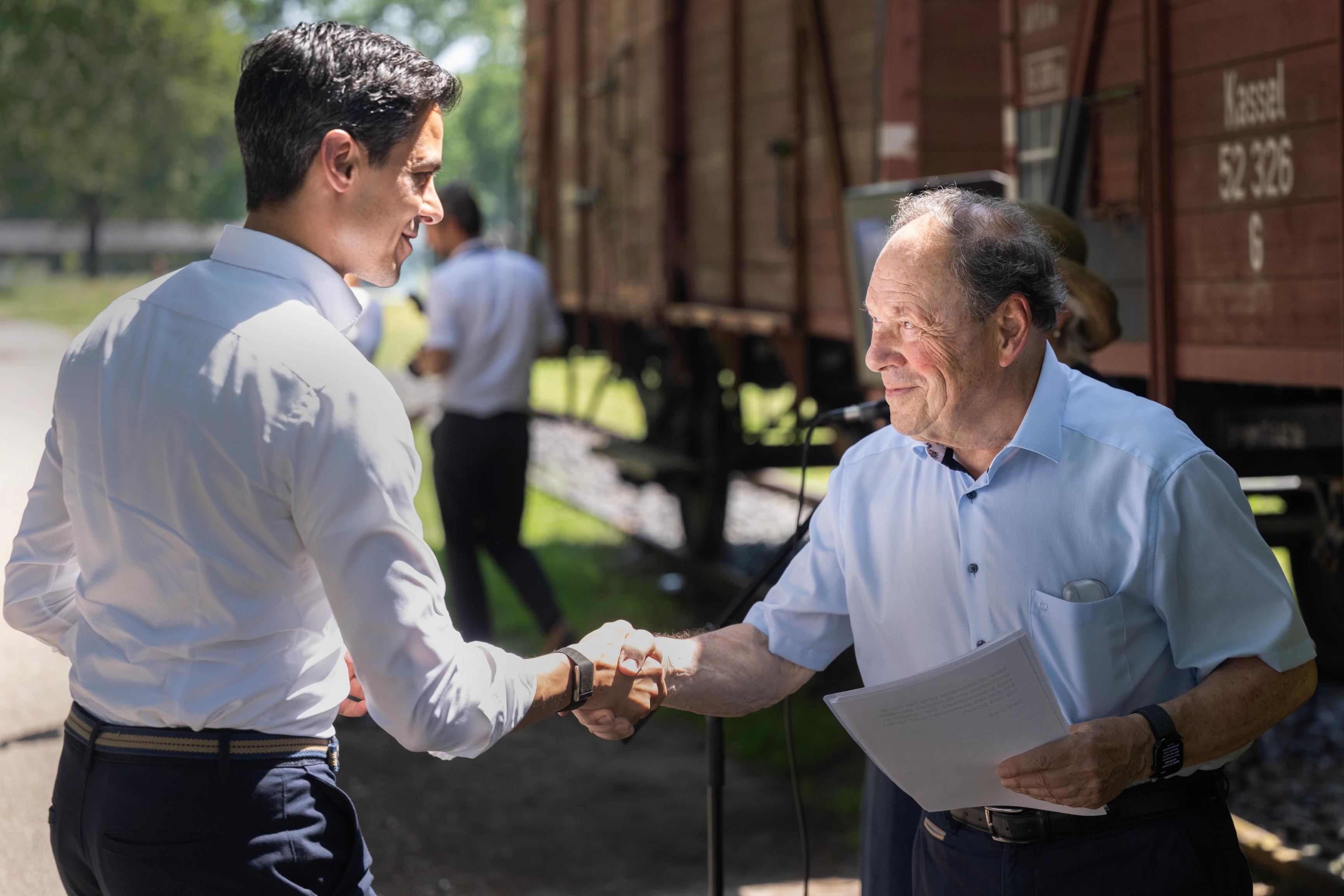 Minister-president Rob Jetten schudt hand met kampoverlevende Hans Peeper voor de wagon op het kampterrein van kamp Westerbork