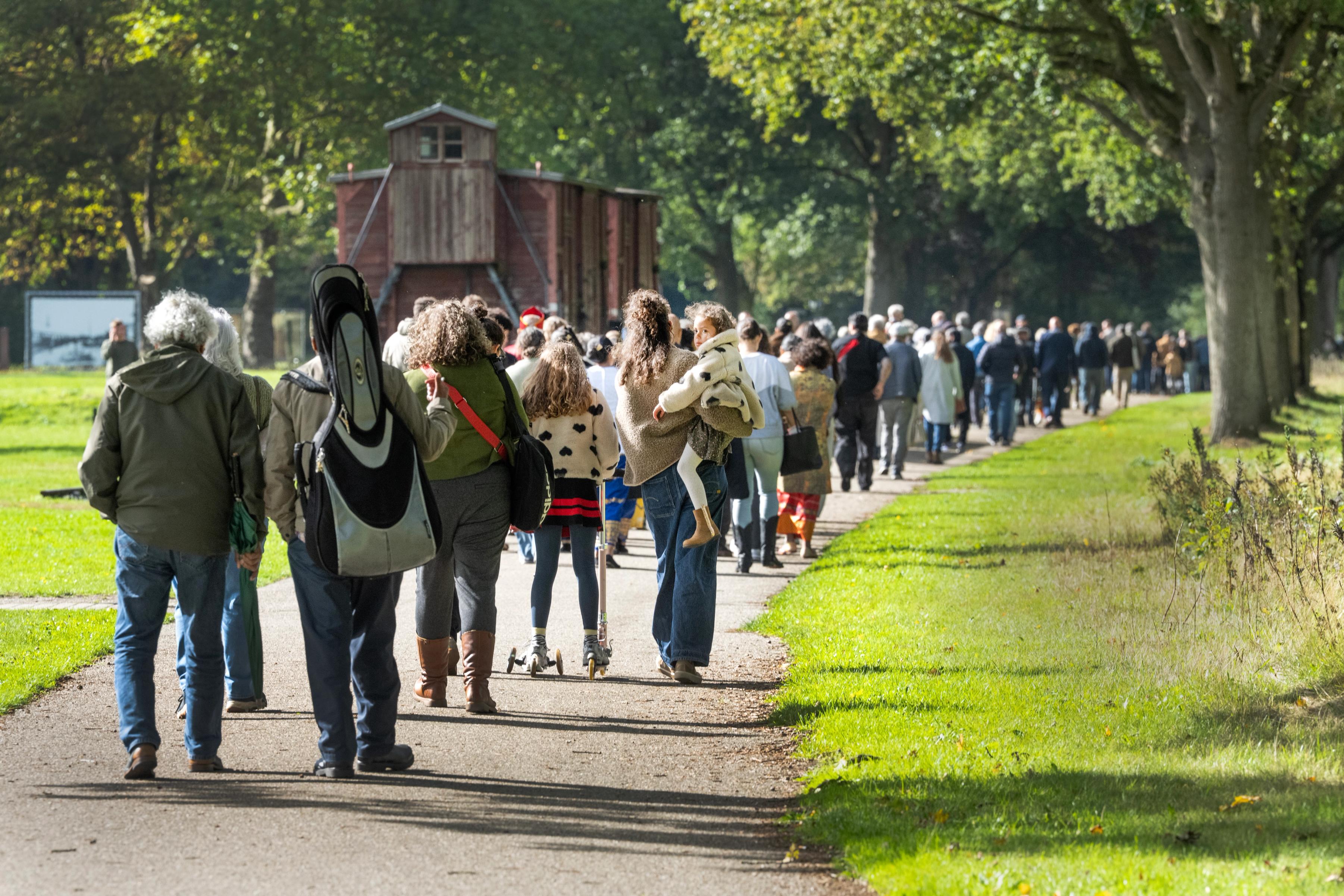 Grote groep bezoekers wandelt over pad op voormalig kampterrein Westerbork langs de treinwagons 