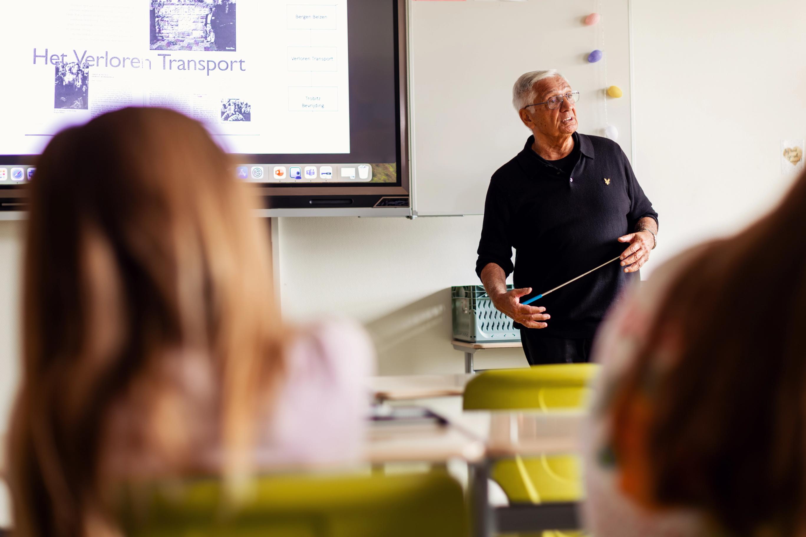 Oudere man met zwarte trui en aanwijsstok in zijn hand staat voor een klas te spreken, met op de voorgrond twee jonge meiden en achter de spreker een beamer met een diavoorstelling over Het Verloren Transport