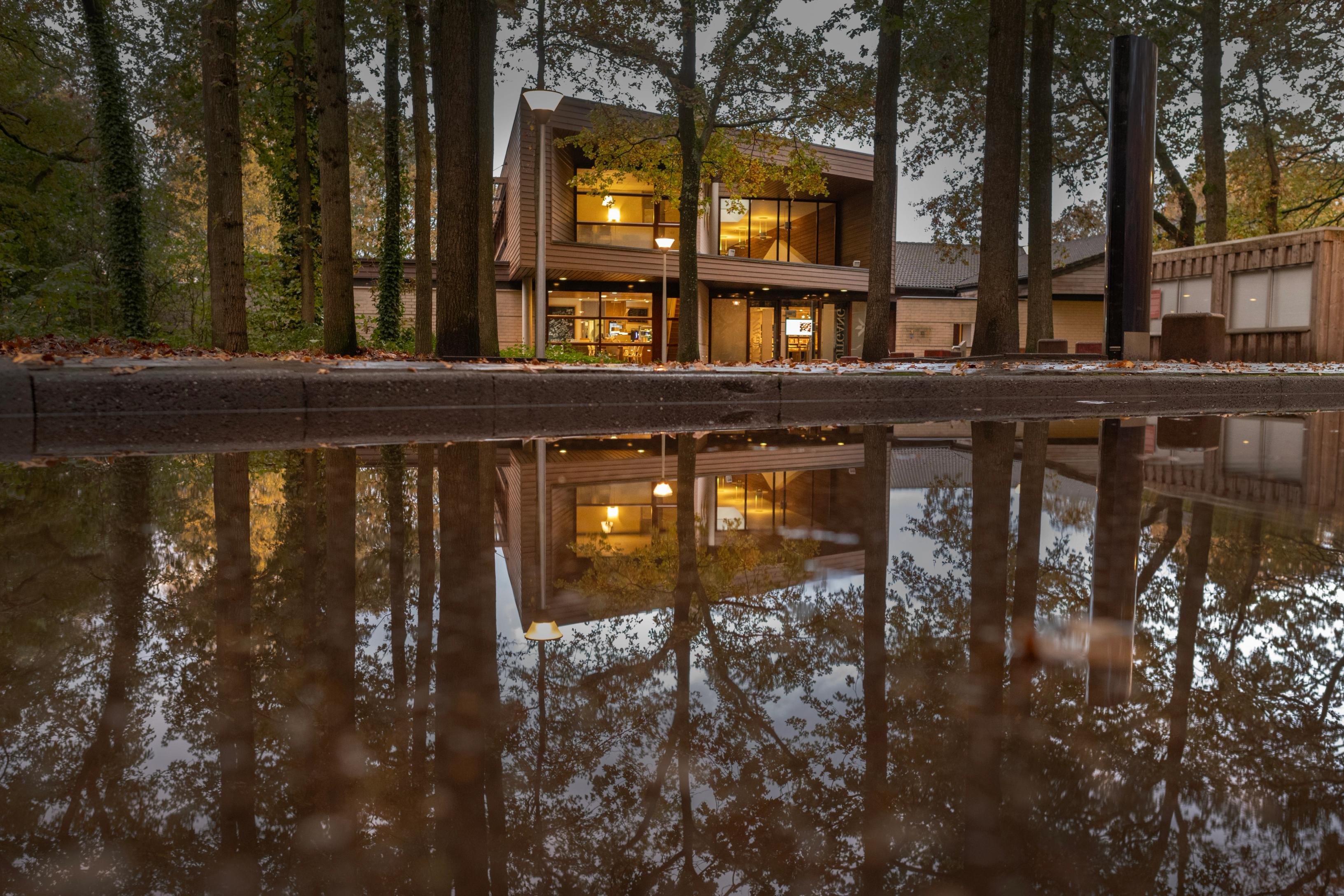 Gebouw van Herinneringscentrum Kamp Westerbork achter een rij bomen met verlichting aan binnen, met op de voorgrond een plas regenwater