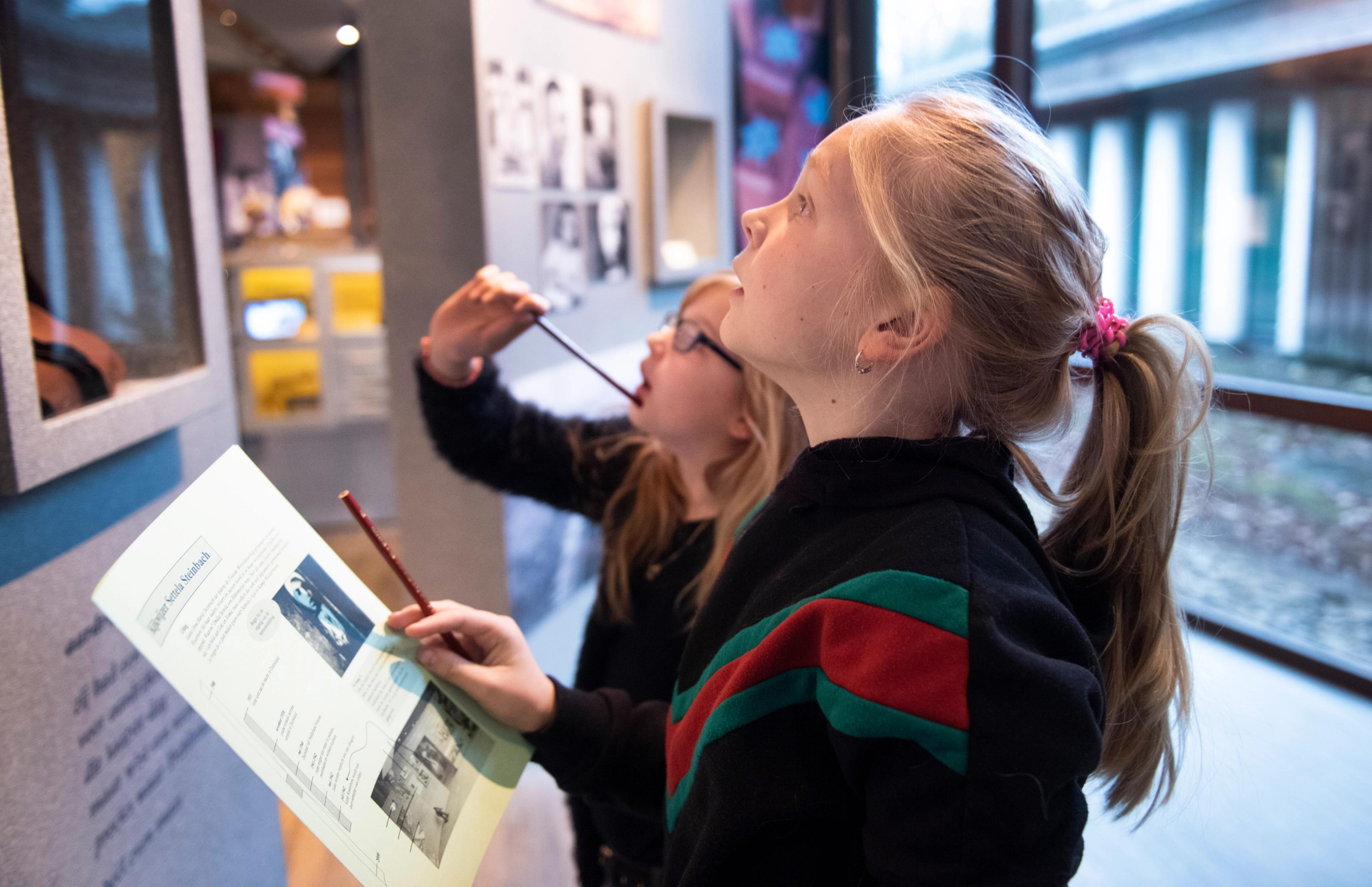 A girl with blonde hair is looking at a wall in the museum for a school assignment, the details of which are explained on a piece of paper she is holding