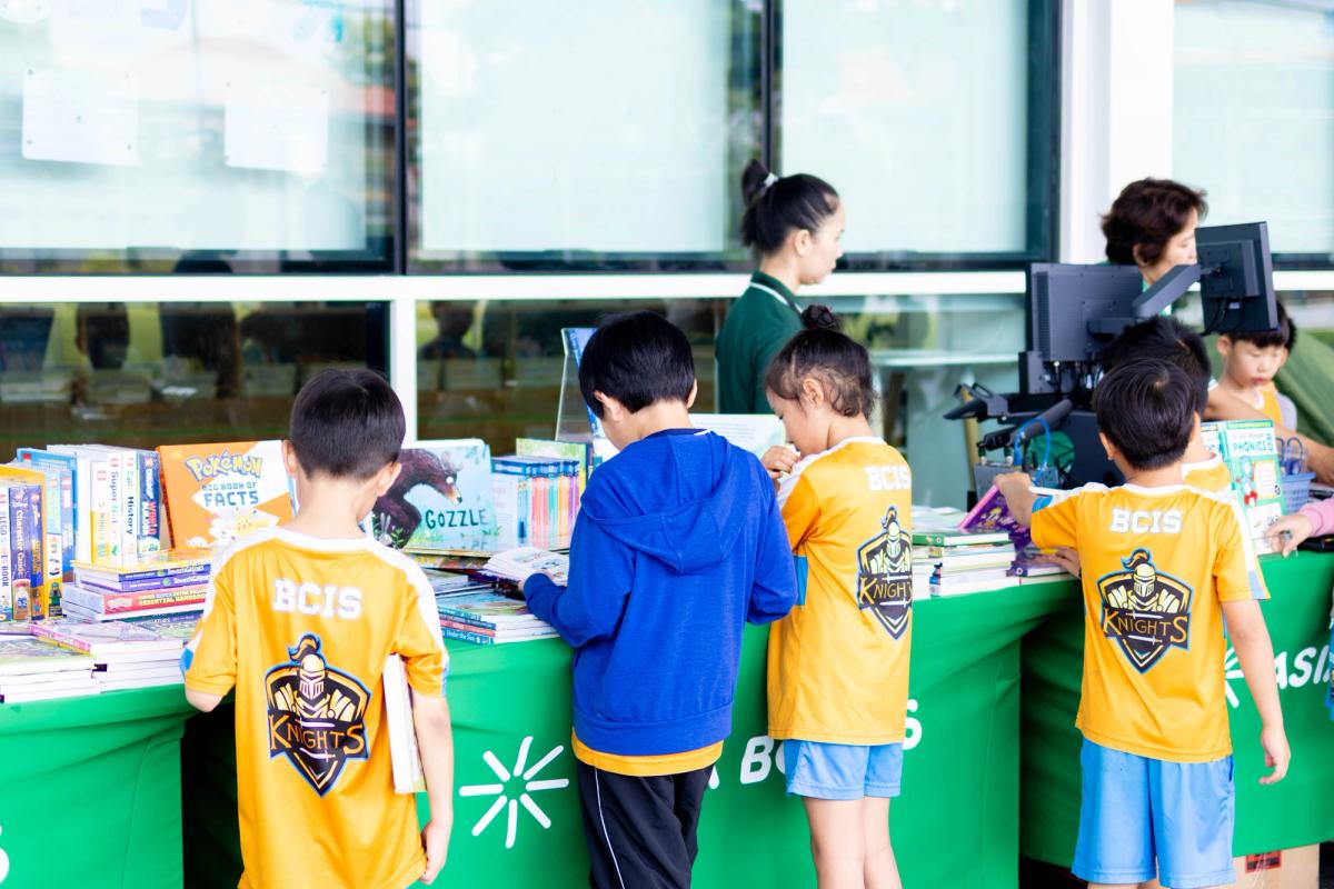 Four BCIS Knights pupils in yellow sports kit browse tables of children's books beneath a green Asia Books banner at the school's Book Fair.