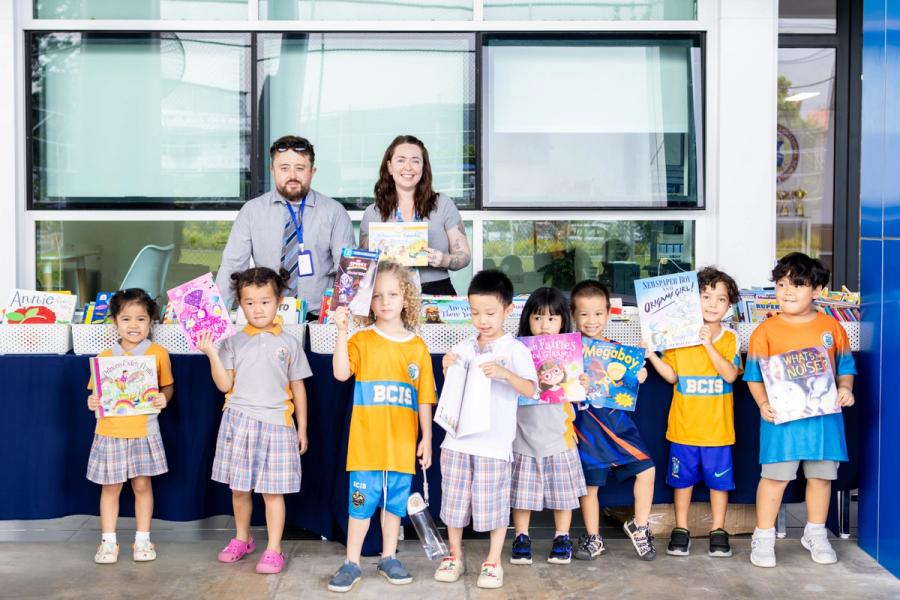 A line of Reception and Year 1 pupils each hold up a book they chose, standing with two staff members in front of the fair's book tables.