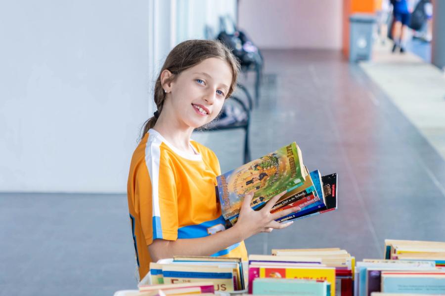 A BCIS pupil in an orange sports shirt smiles while holding a stack of chosen books at the fair's display tables.