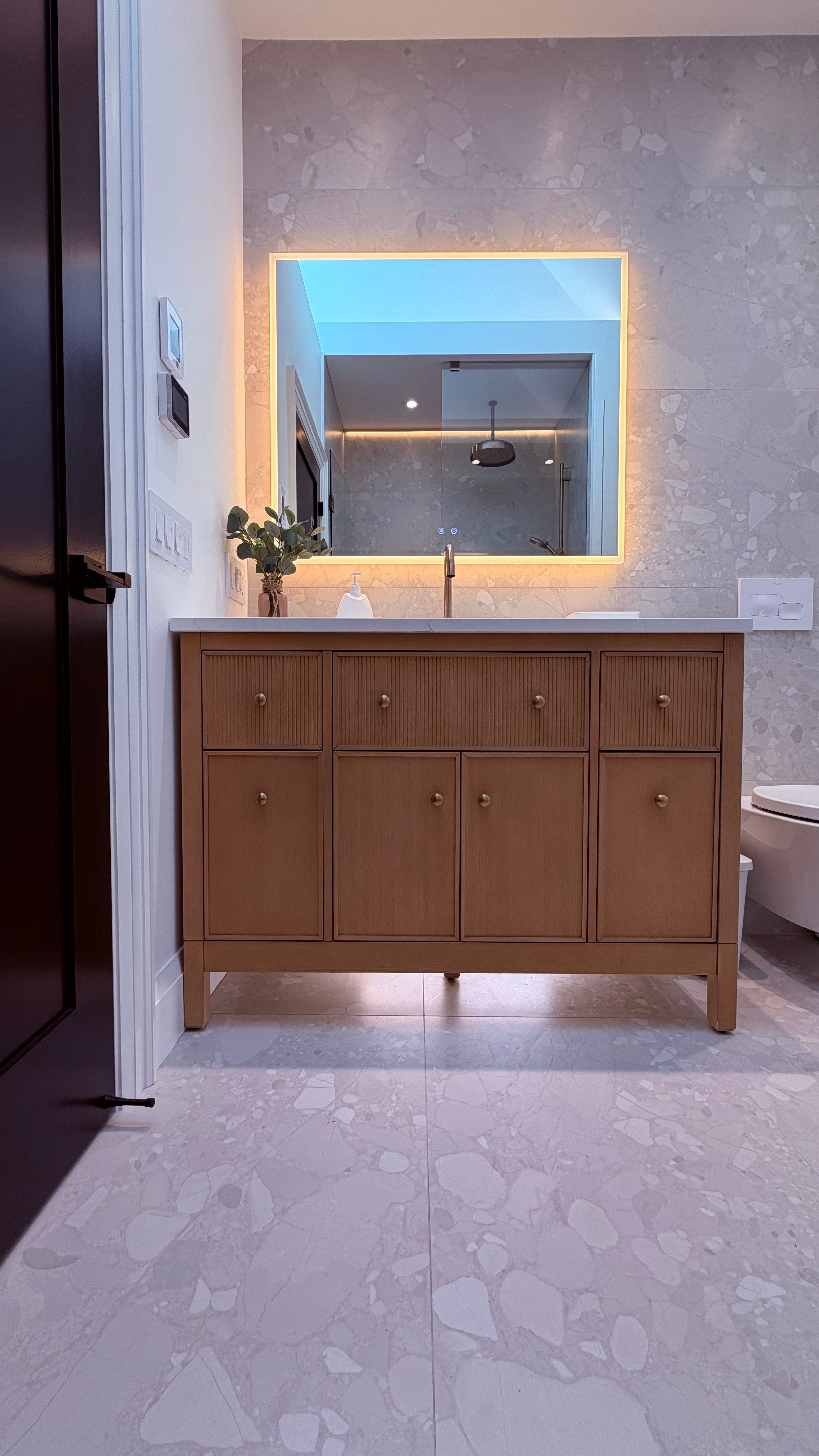 A renovated bathroom with a wall-hung toilet, a backlit mirror, under-cabinet lighting, and heated flooring.