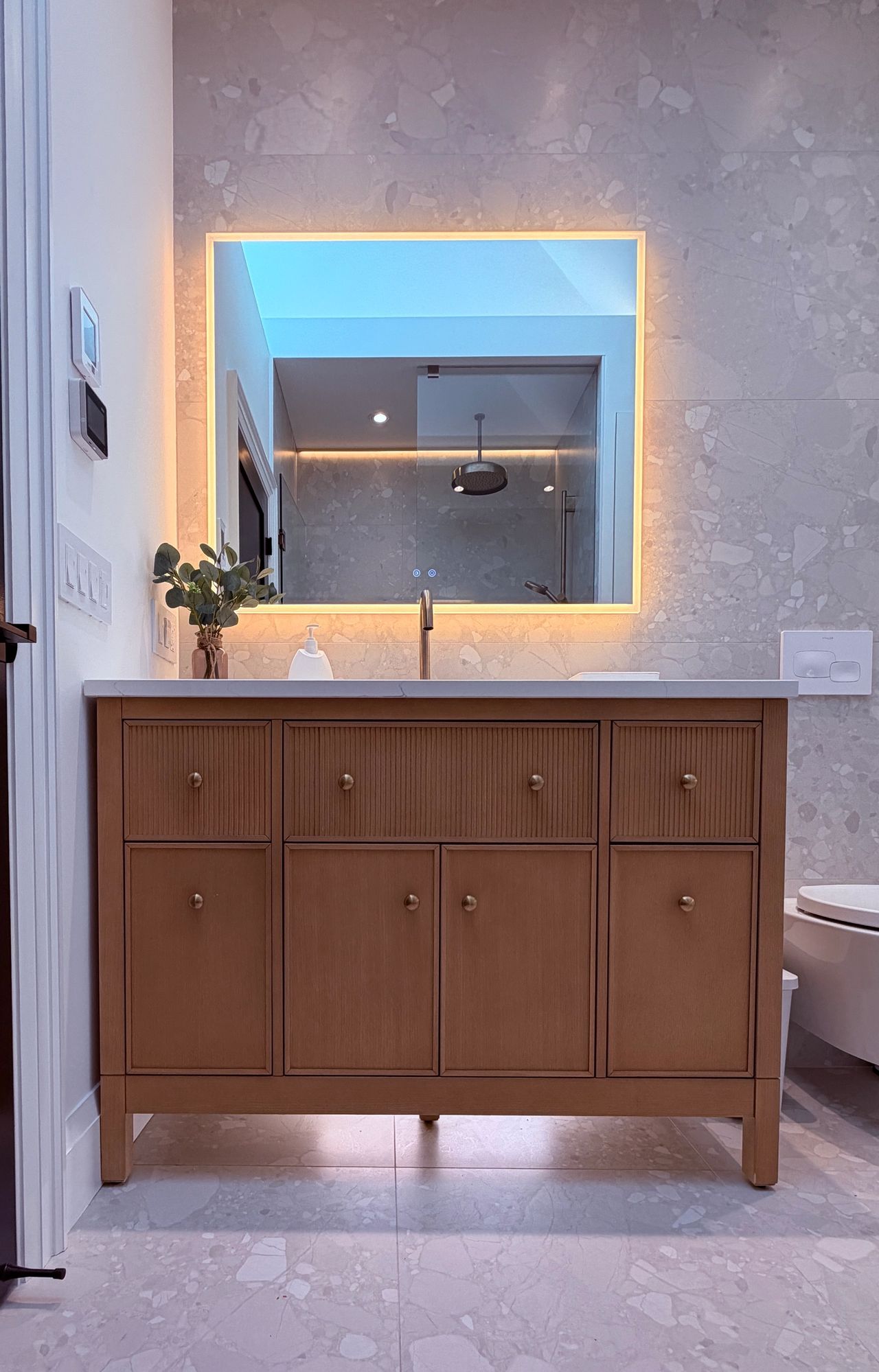 A renovated bathroom with a wall-hung toilet, a backlit mirror, under-cabinet lighting, and heated flooring.