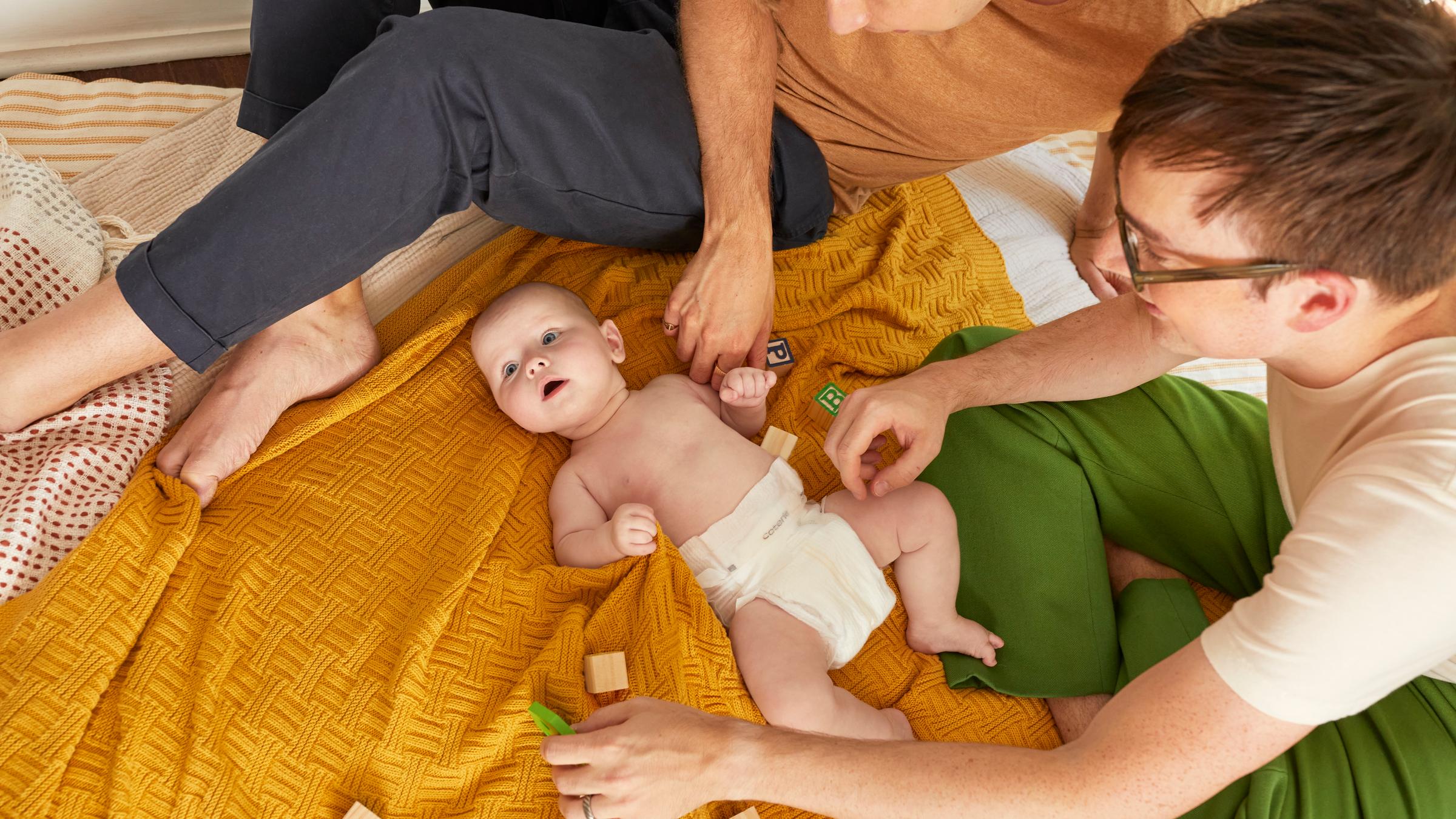 Two dads are playing with their child who is laying on a yellow blanket.