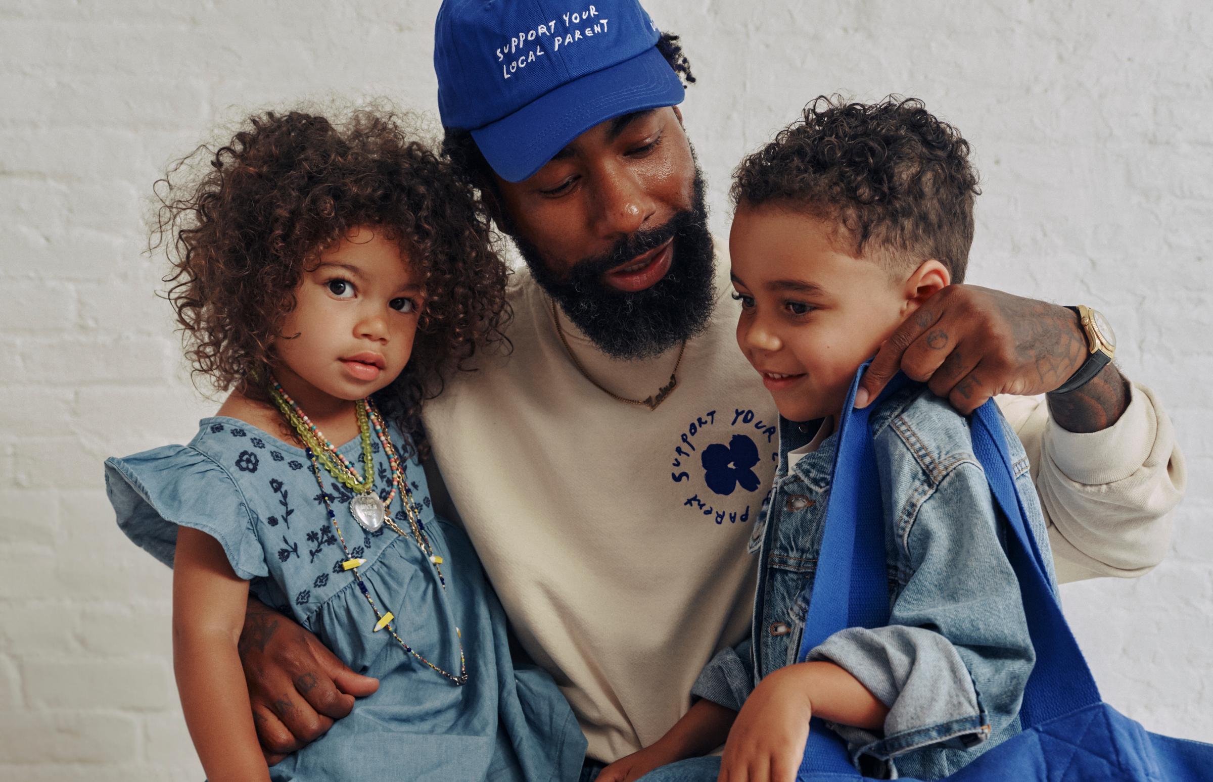 A father holds his two kids while wearing the Support Your Local Parent crewneck and hat.