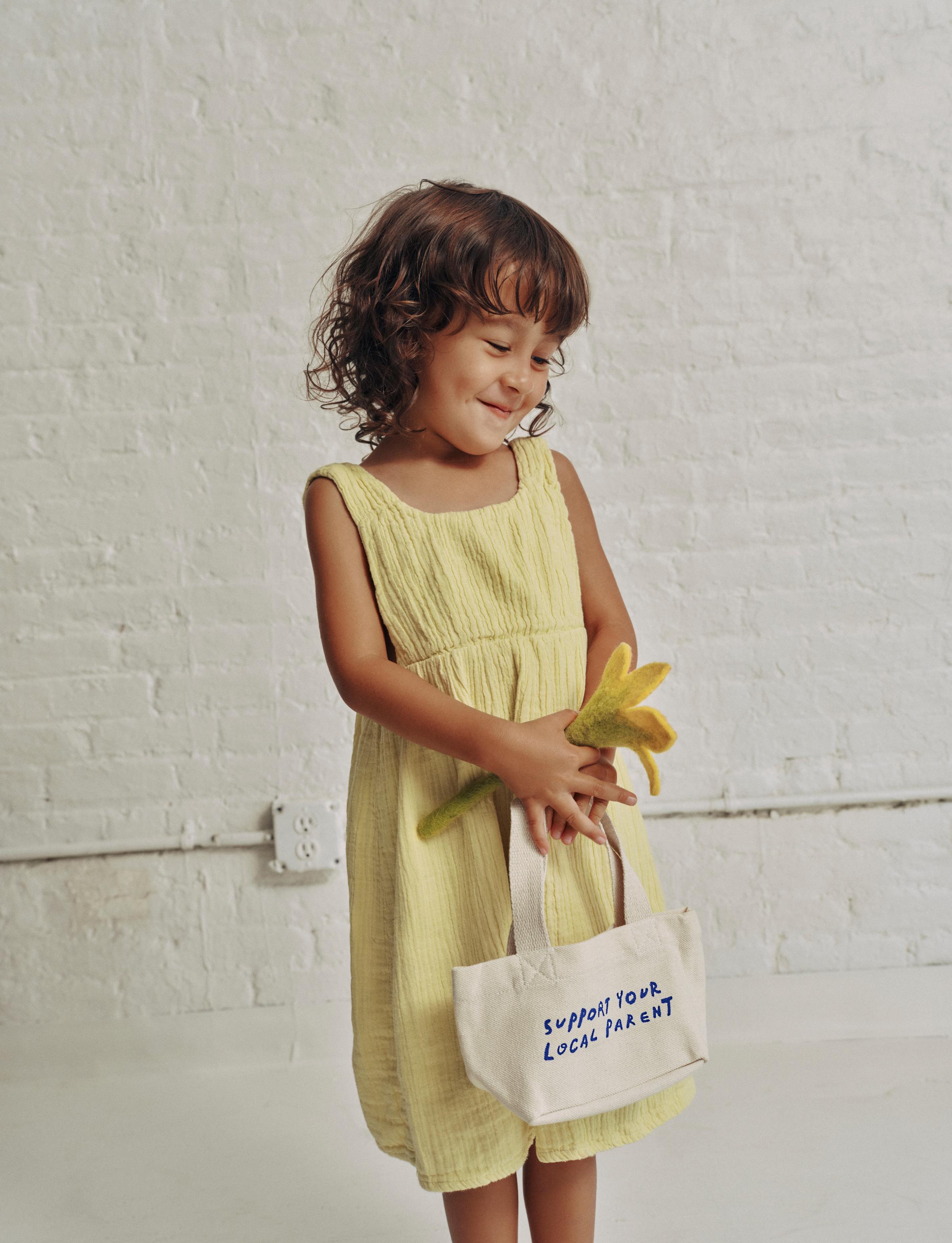 A little girl in a yellow dress holds a fabric toy flower and the mini SYLP tote.