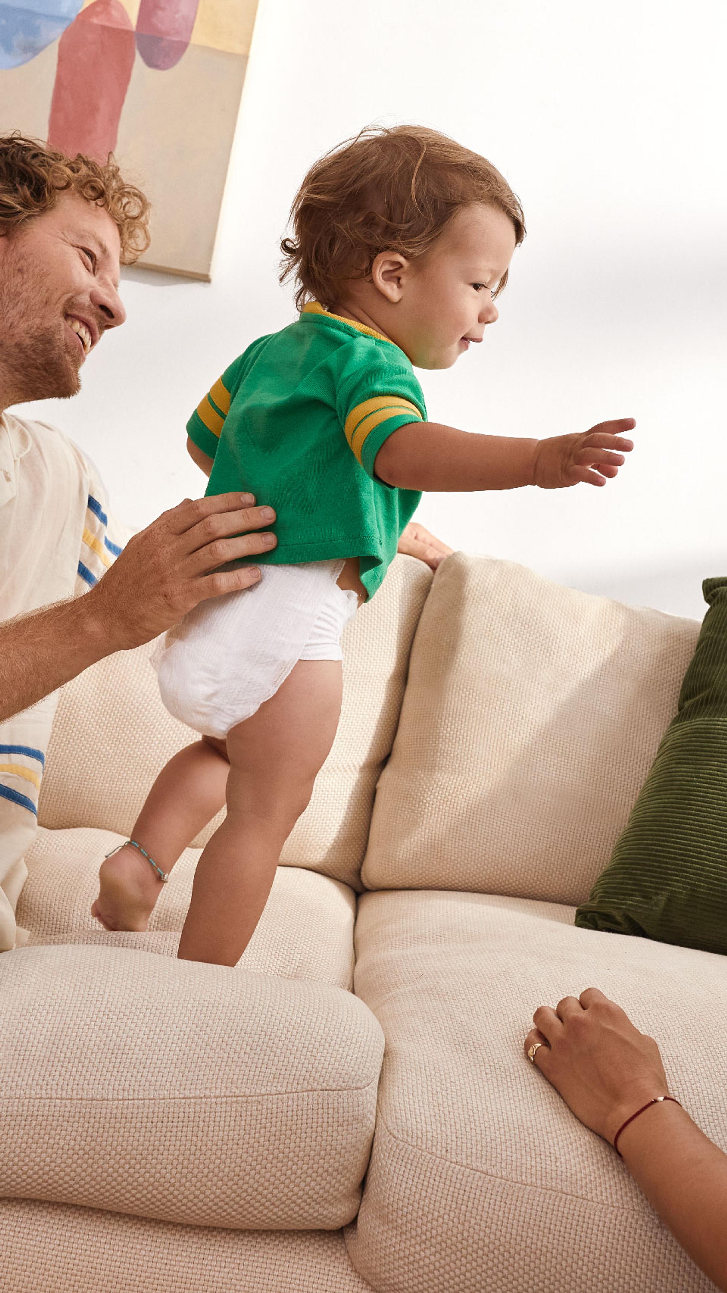 a baby stands up on a couch with the support of his mother and father.
