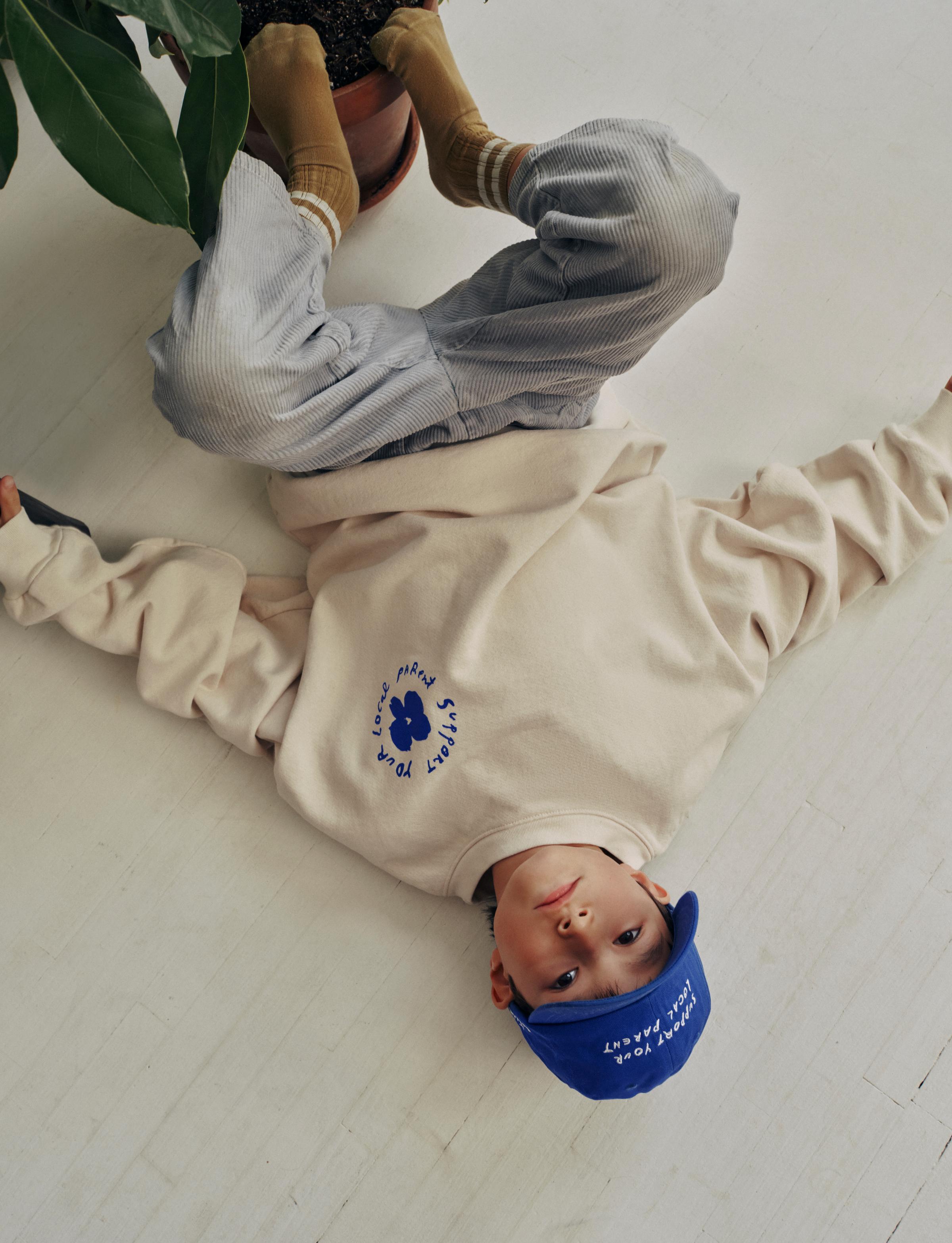 A child lays on the floor in an oversized SYLP crewneck and electric blue hat.