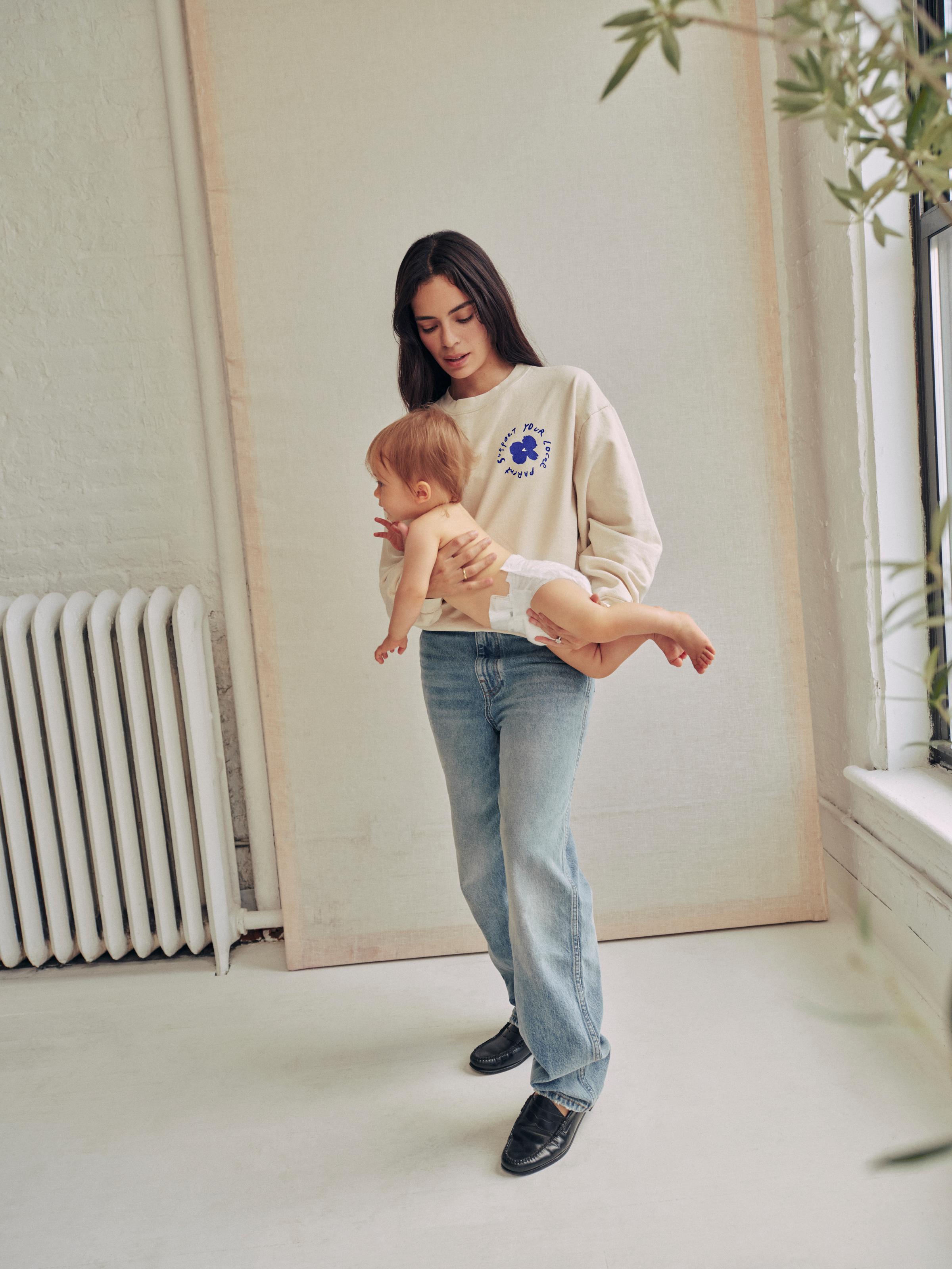 A woman is holding a baby while wearing the SYLP crewneck.