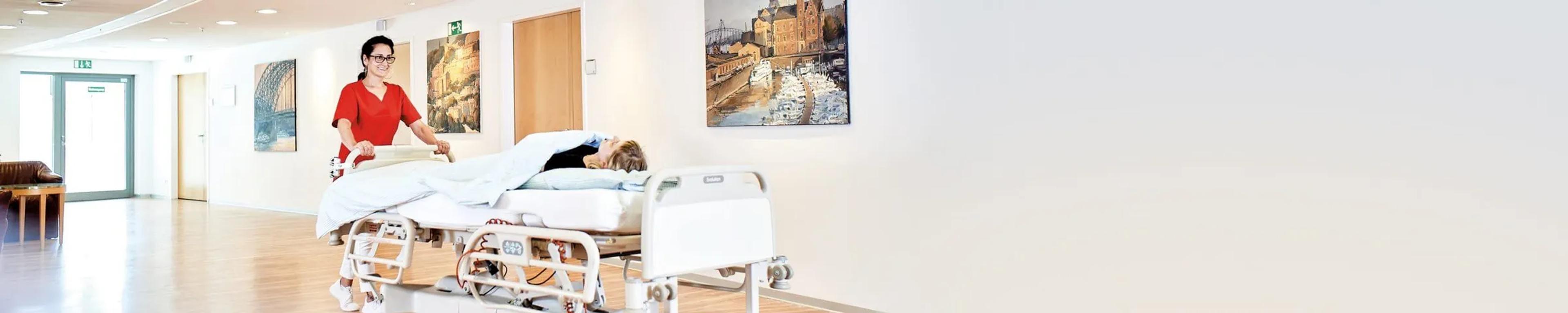 Eine medizinische Fachangestellte schiebt eine Patientin der Orthopädie Mediapark in ihrem Bett durch die Klinik.