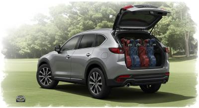 ゴルフ場の駐車エリアで停車するヴォクシー。開いたテールゲート付近に4つのゴルフバッグが見え、余裕ある積載感を示す。中〜遠景、自然光で落ち着いたクールトーン。ロゴは画像の端に小さく配置（車・バッグ上は不可）。