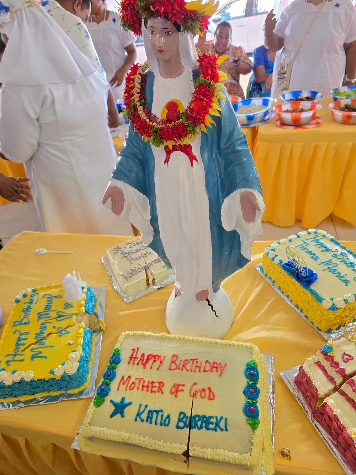 A statue of Mother Mary adorned with a flower lei and surrounded by birthday cakes