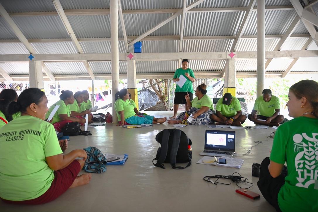 A woman standing and speaking while a group of people seated on the floor look up at her