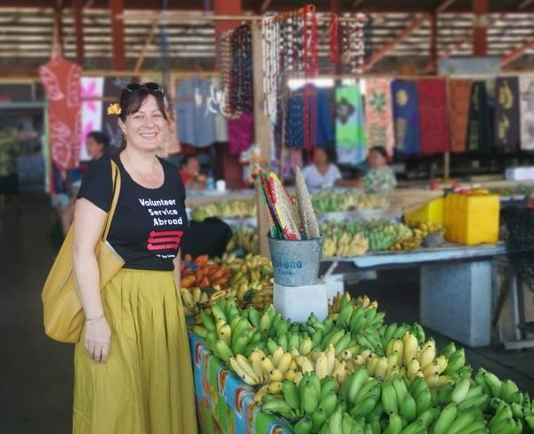 A woman smiles and stands next to a fruit stall at a market
