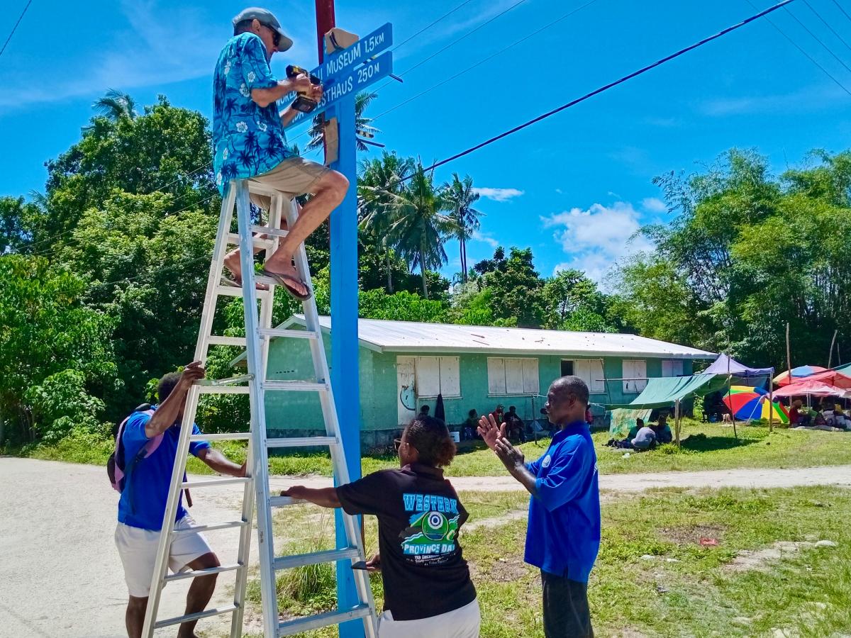 A man sits on the top of a ladder drilling a sign, while three people hold the bottom of the ladder