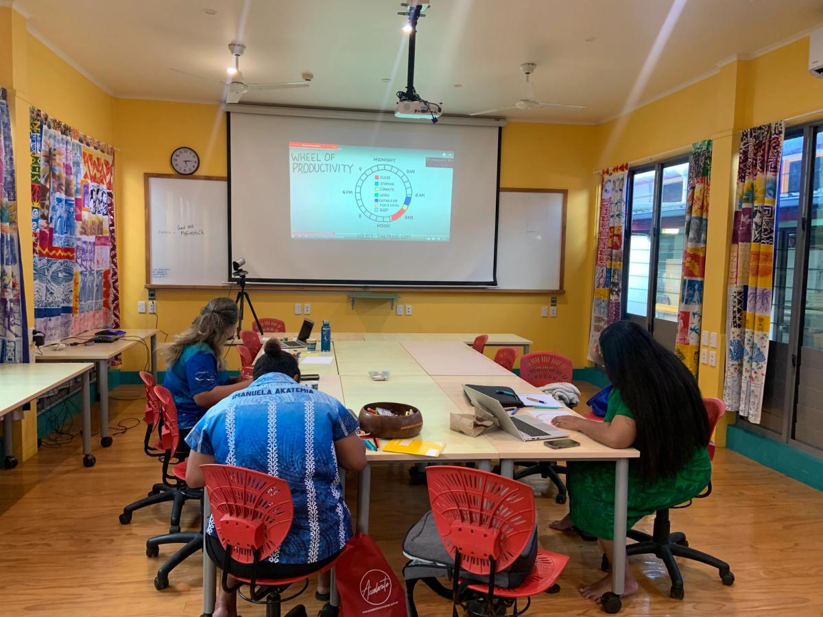 Three students sit around a table located in front of a projector screen inside of a classroom. Three students sit around a table located in front of a projector screen inside of a classroom.